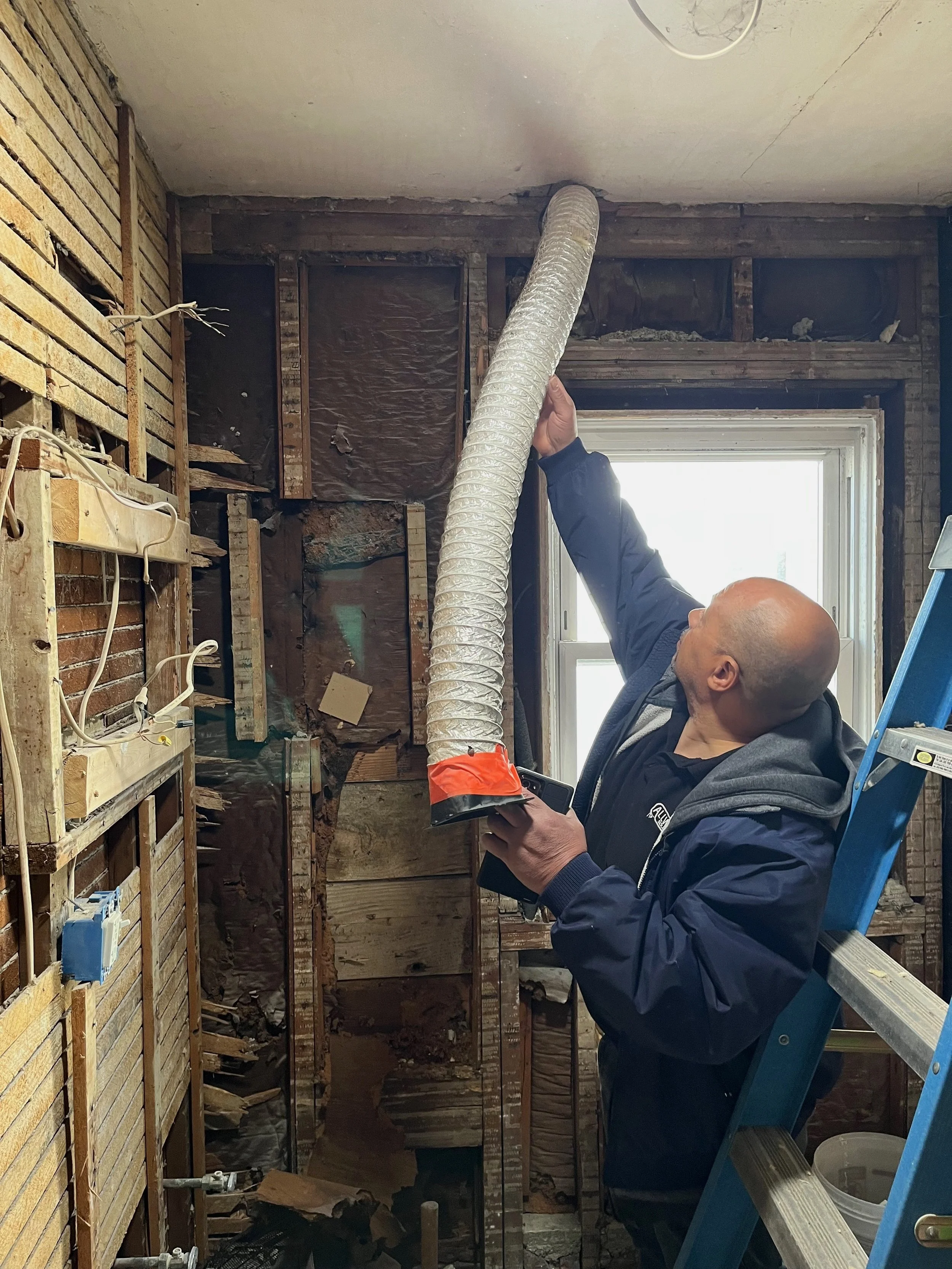 Electrician standing on a ladder is holding a flexible ventilation duct in a room undergoing renovation with exposed wall framing and an open window.