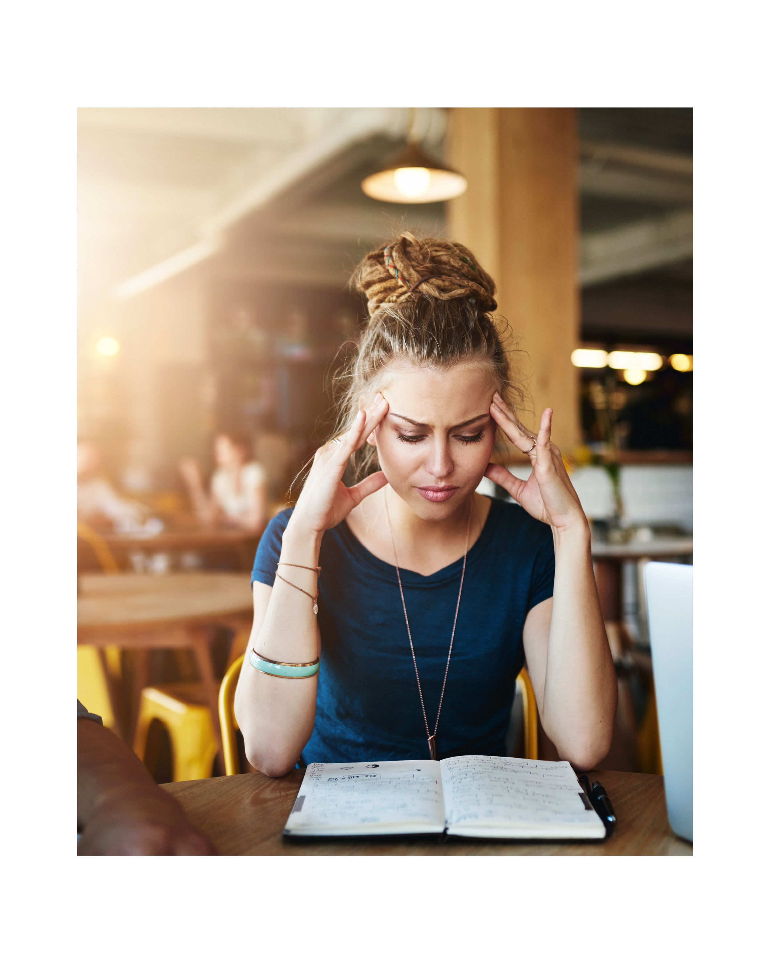 A woman with dreadlocks and wearing a blue shirt sitting at a table in a coffee shop, holding her head with both hands, appearing stressed or concentrated while looking at an open notebook.