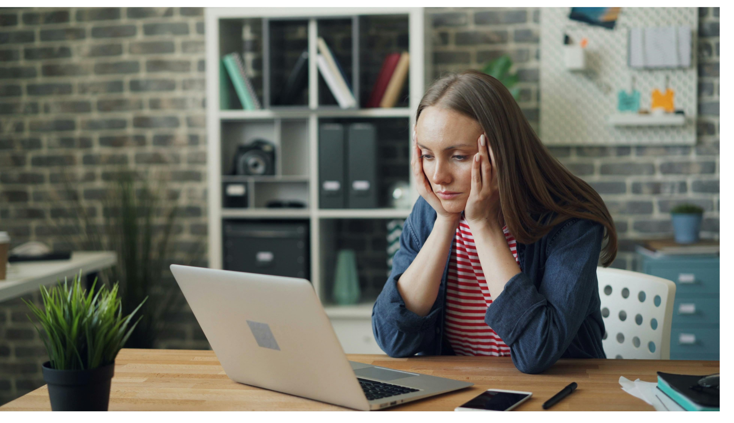 Young woman sitting at a desk in front of a laptop computer, looking stressed or exhausted, with her hands on her head. There are potted plants, a smartphone, a pen, and notebooks on the desk, and background features a brick wall, bookshelf, and bulletin board.