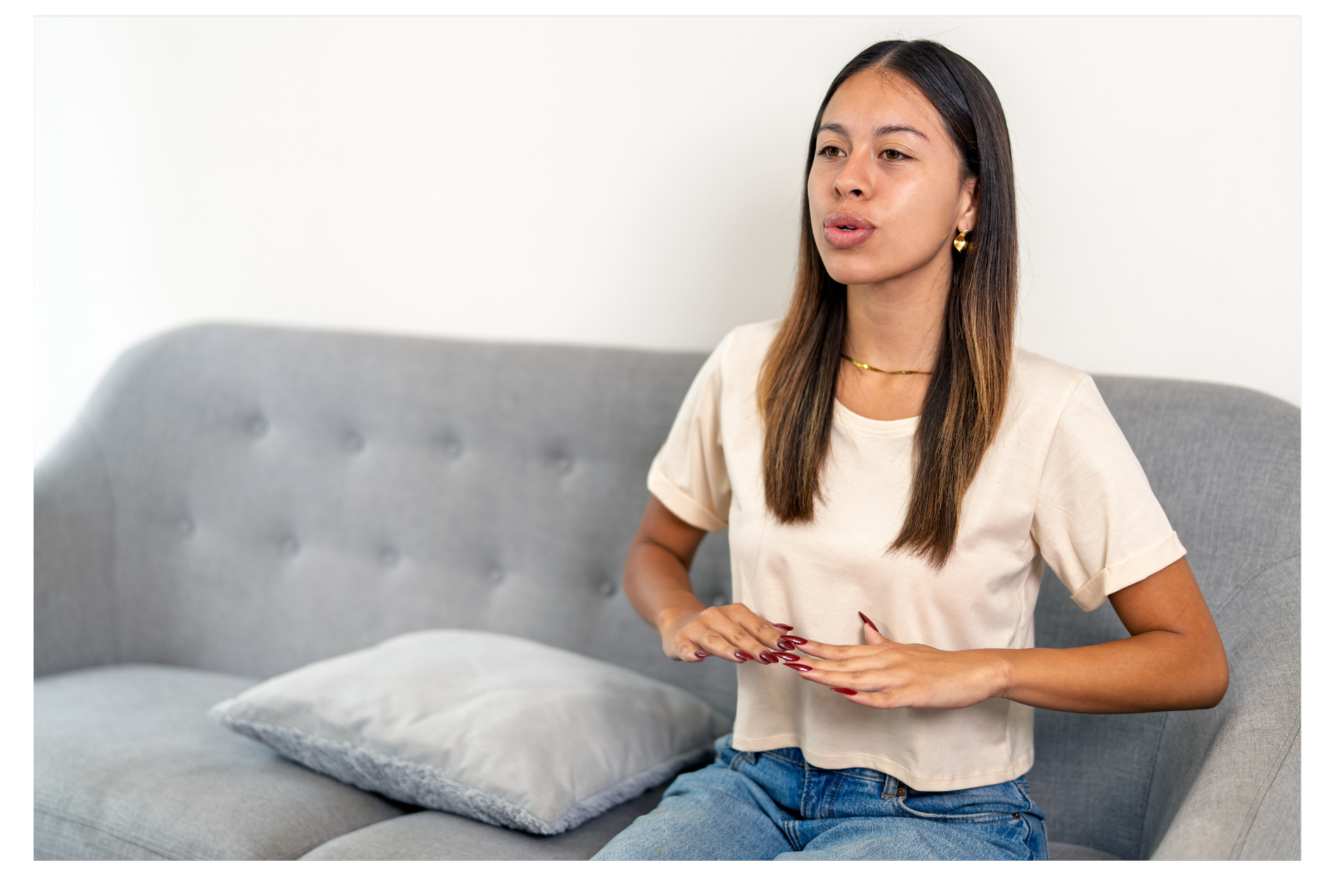 A woman with long brown hair, wearing a beige t-shirt and blue jeans, speaking and gesturing with her hands while sitting on a gray couch with a pillow.