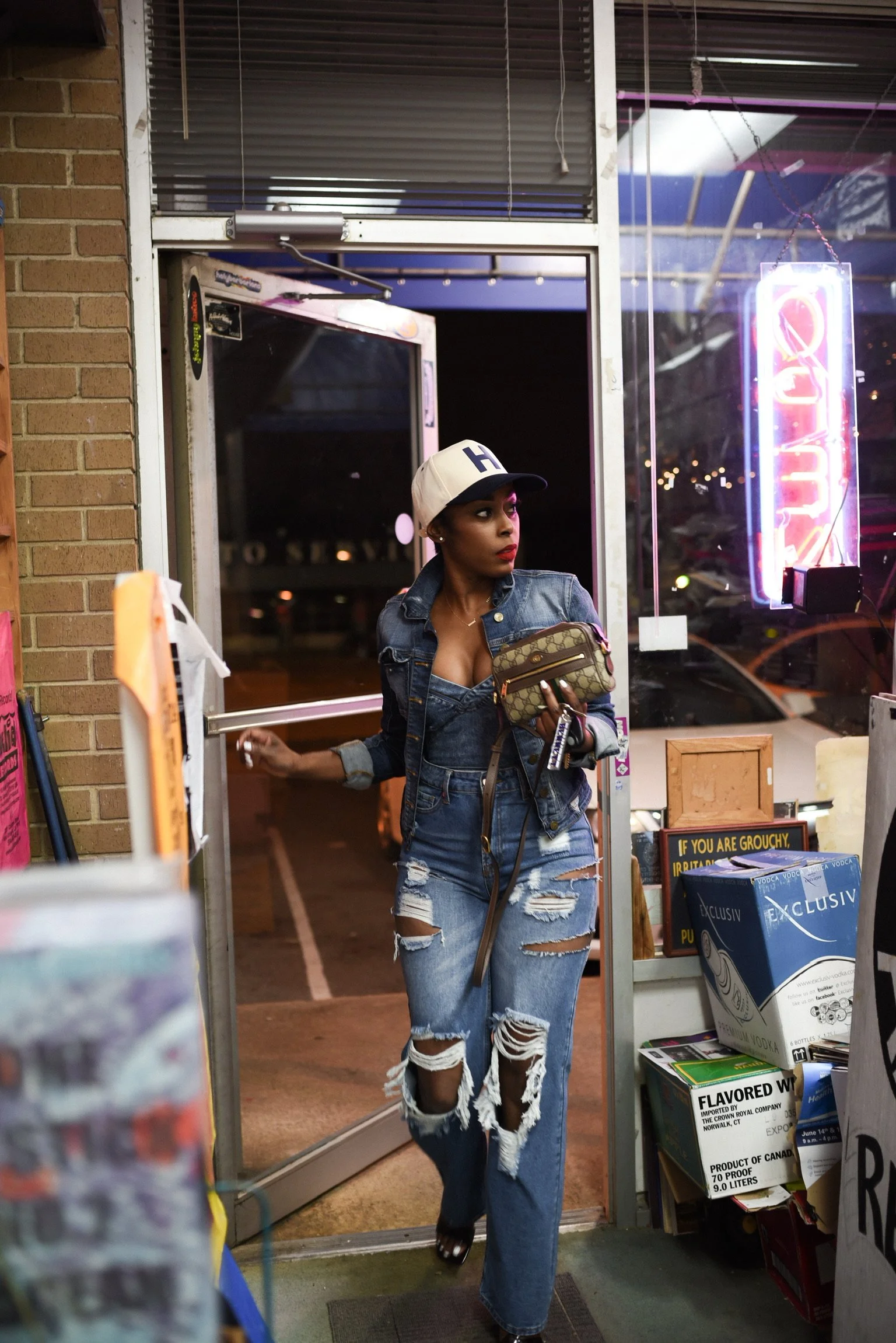 A person wearing a denim jacket, ripped jeans, and a baseball cap entering a store with neon signs and boxes.