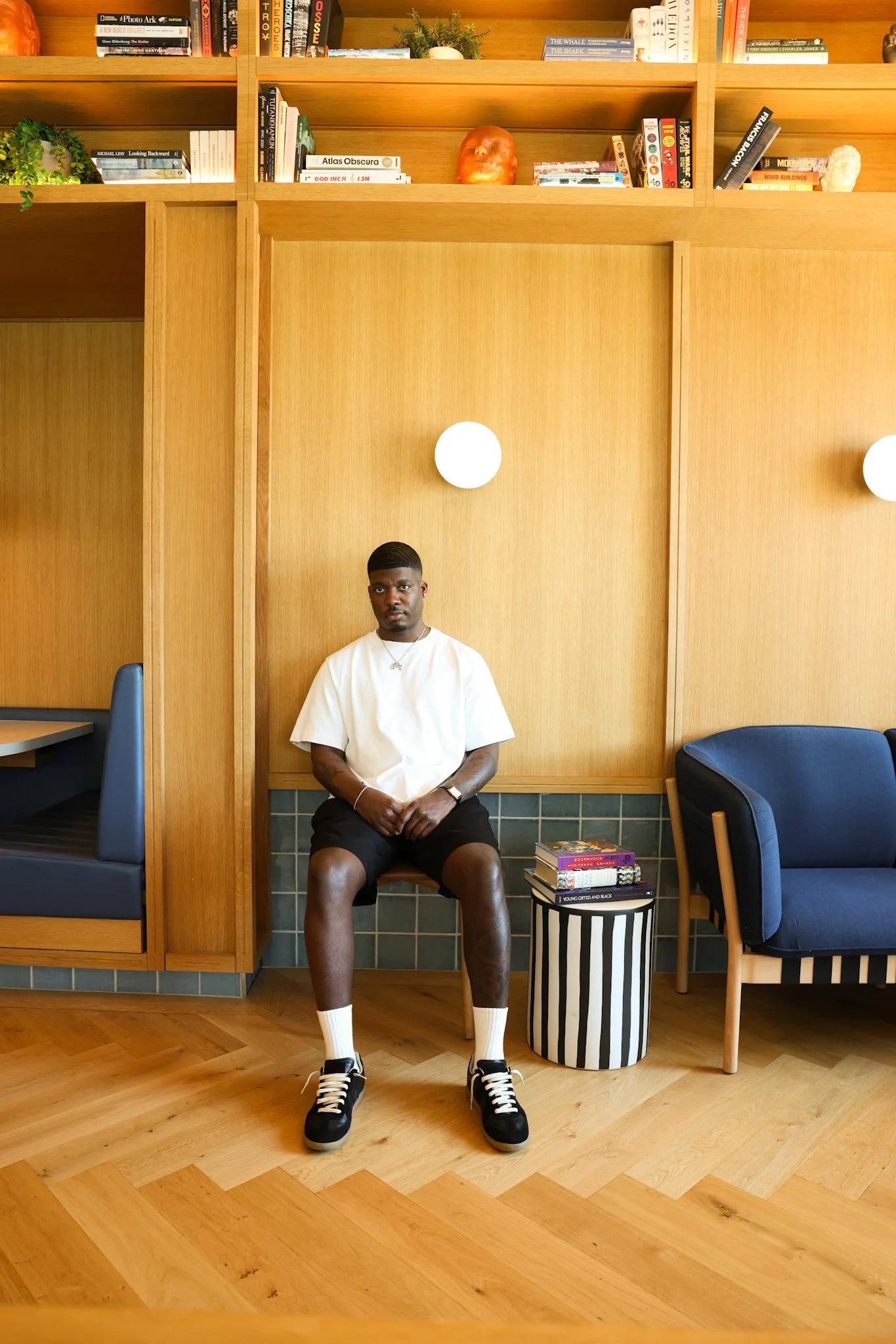 Person sitting on a chair in a modern library setting with wooden interior, surrounded by books on shelves, wearing a white shirt and black shorts.