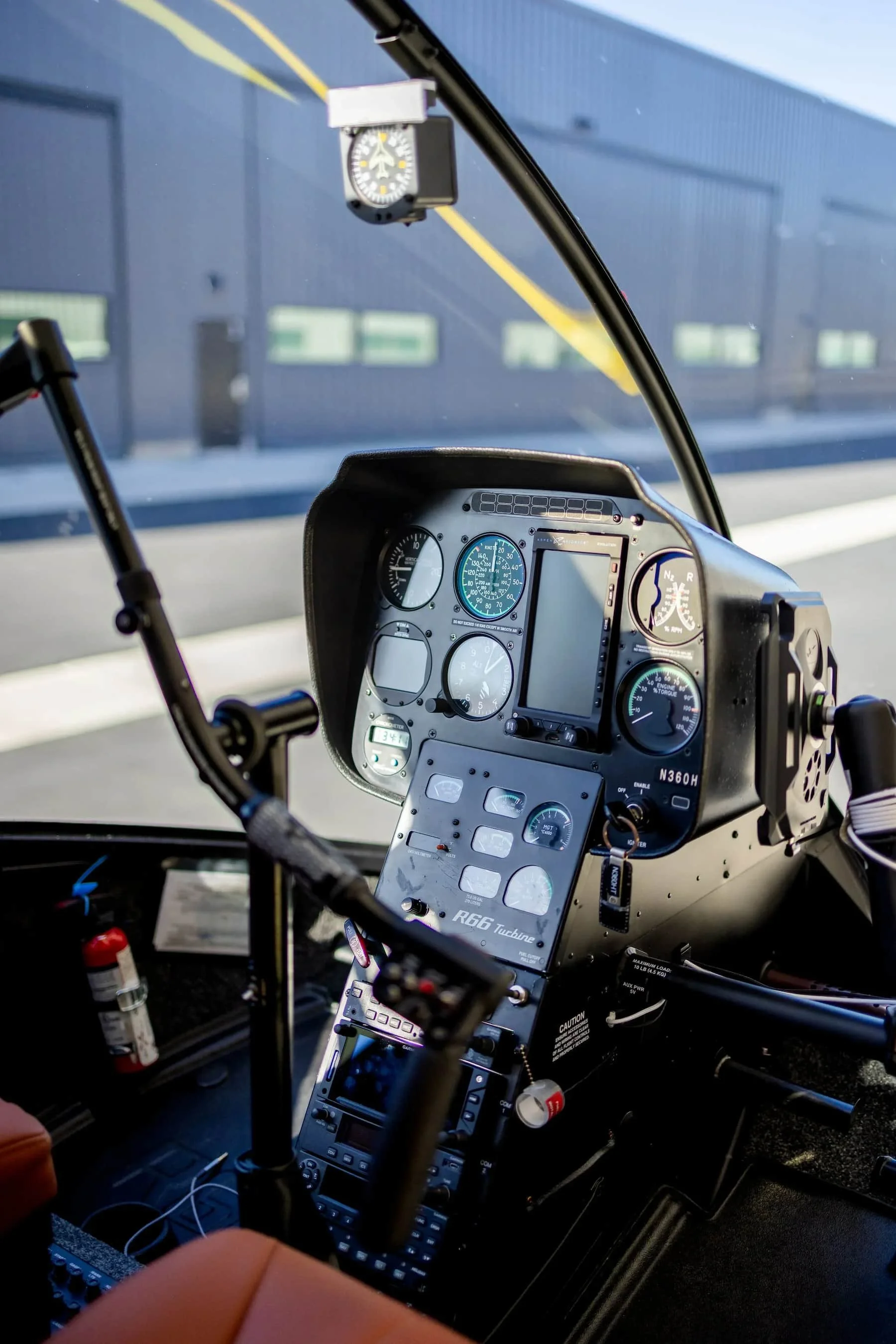 Inside the cockpit of a helicopter, showing various control panels, gauges, and instruments. There is a digital display, rotary switches, and a stick grip in the foreground. The background includes a window revealing an exterior blue building with yellow lines.