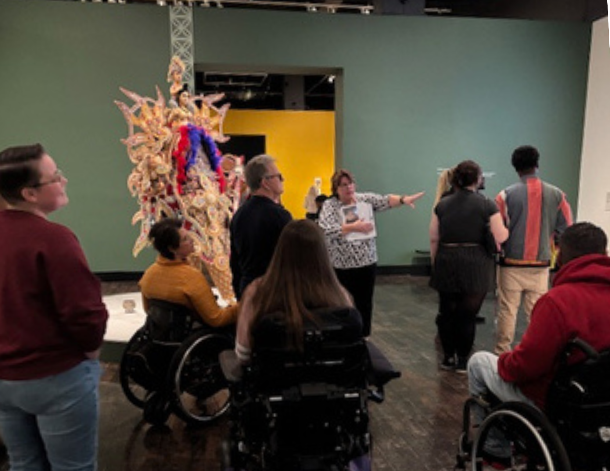 A docent gives a tour of an art museum. Several patrons are listening including 3 in wheelchairs.