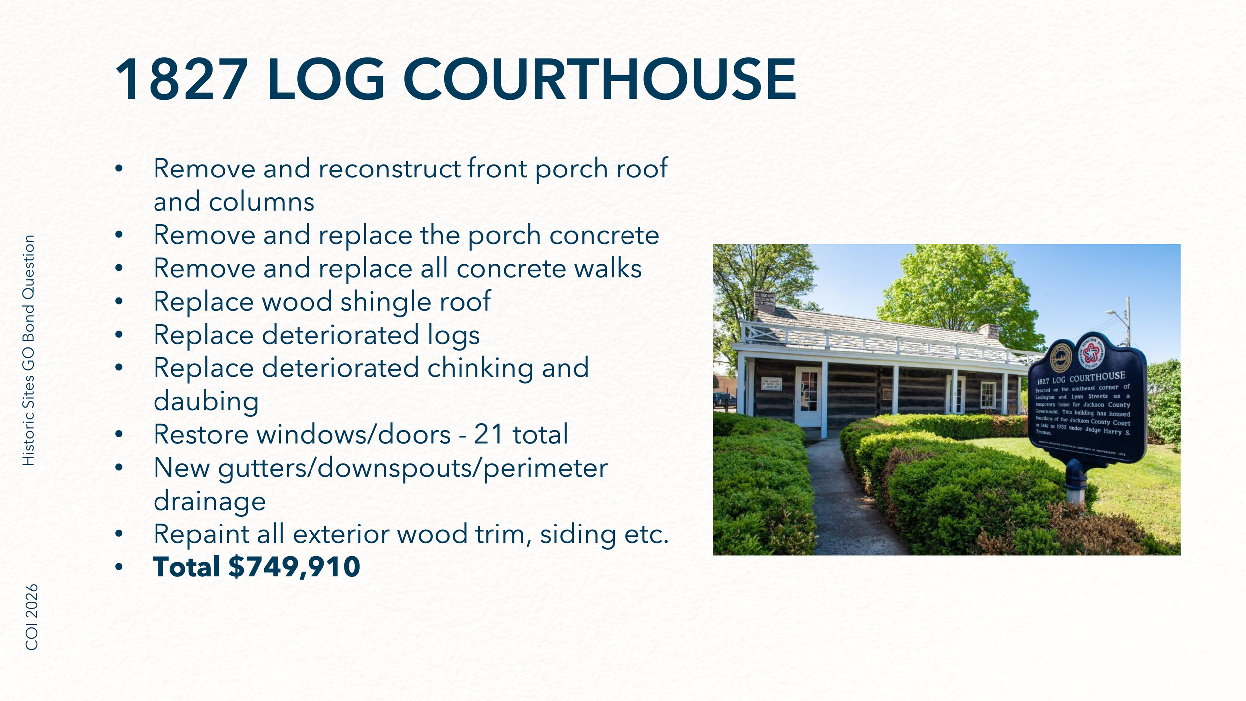 Photo of the 1827 Log Courthouse with a blue sign in front of it that provides historical information. The building is a small, two-story log structure with white trim, surrounded by green bushes and trees. The sky is clear and blue.