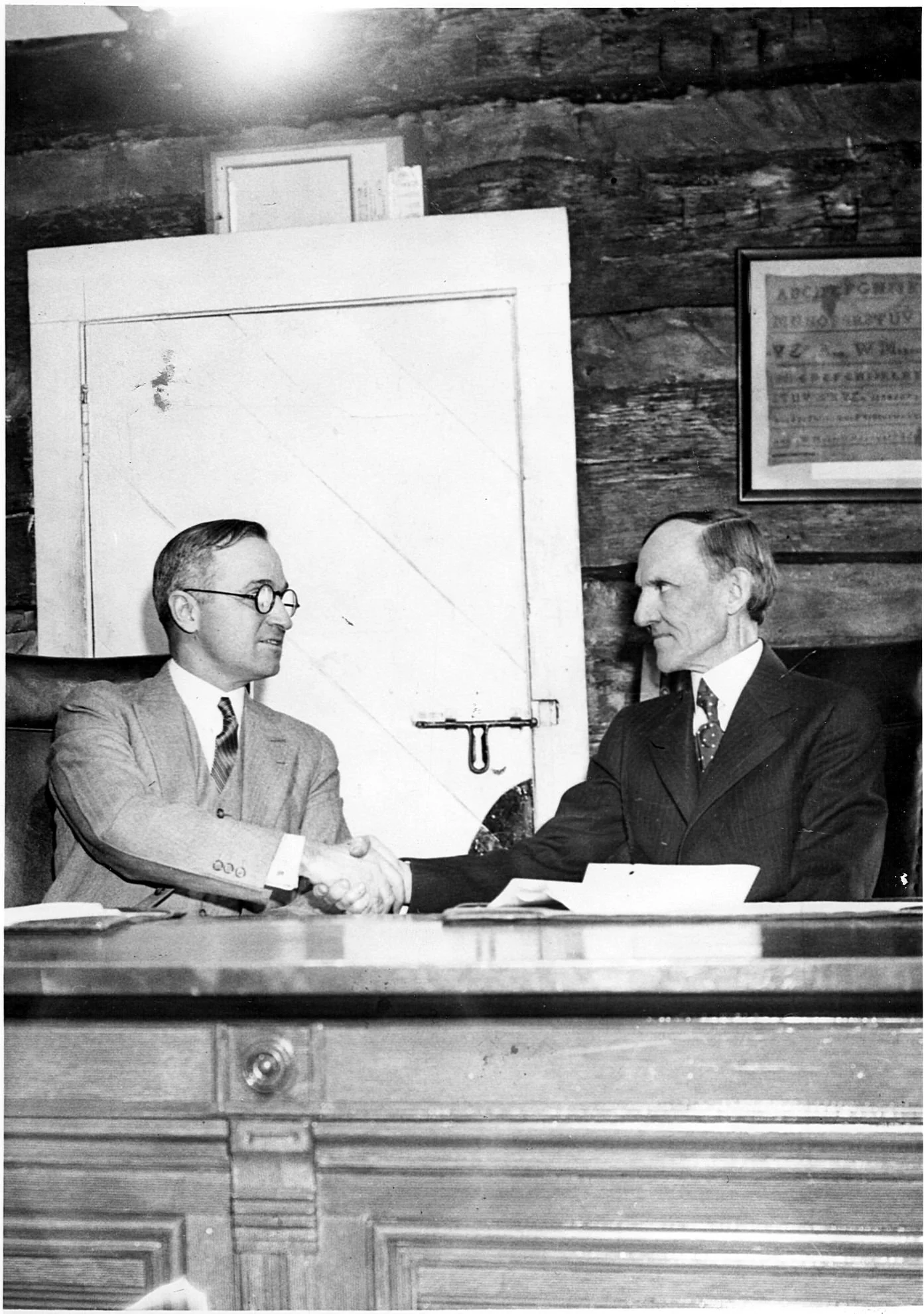 Black and white photo showing two men in suits shaking hands and smiling during a formal meeting or treaty signing, seated behind a desk in a wood-paneled room.