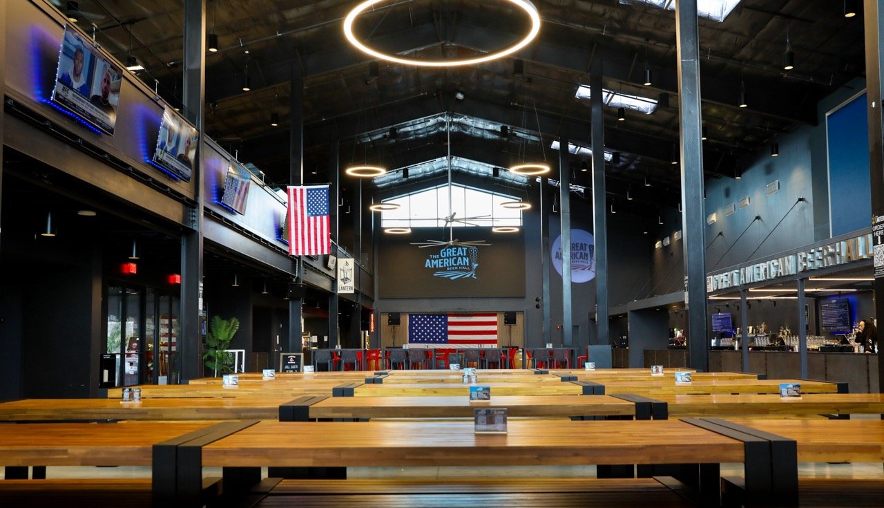Interior of a modern sports bar or event space with high ceilings, circular hanging lights, wooden tables, and American flags. Large screens display sports or news, and there is a bar area on the right side.