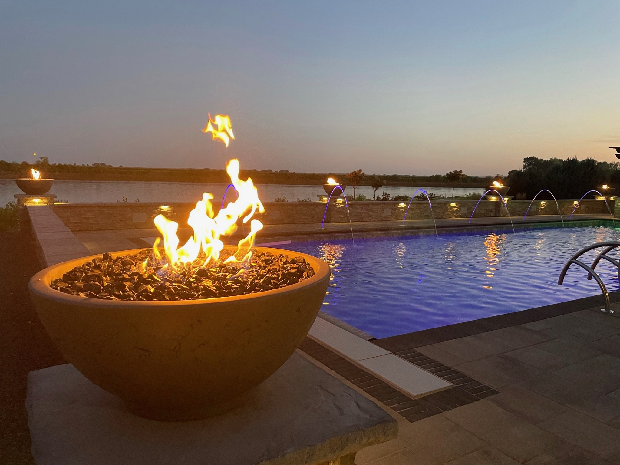 A lit fire in a stone bowl near a swimming pool during dusk with a sunset sky and fountains in the background.