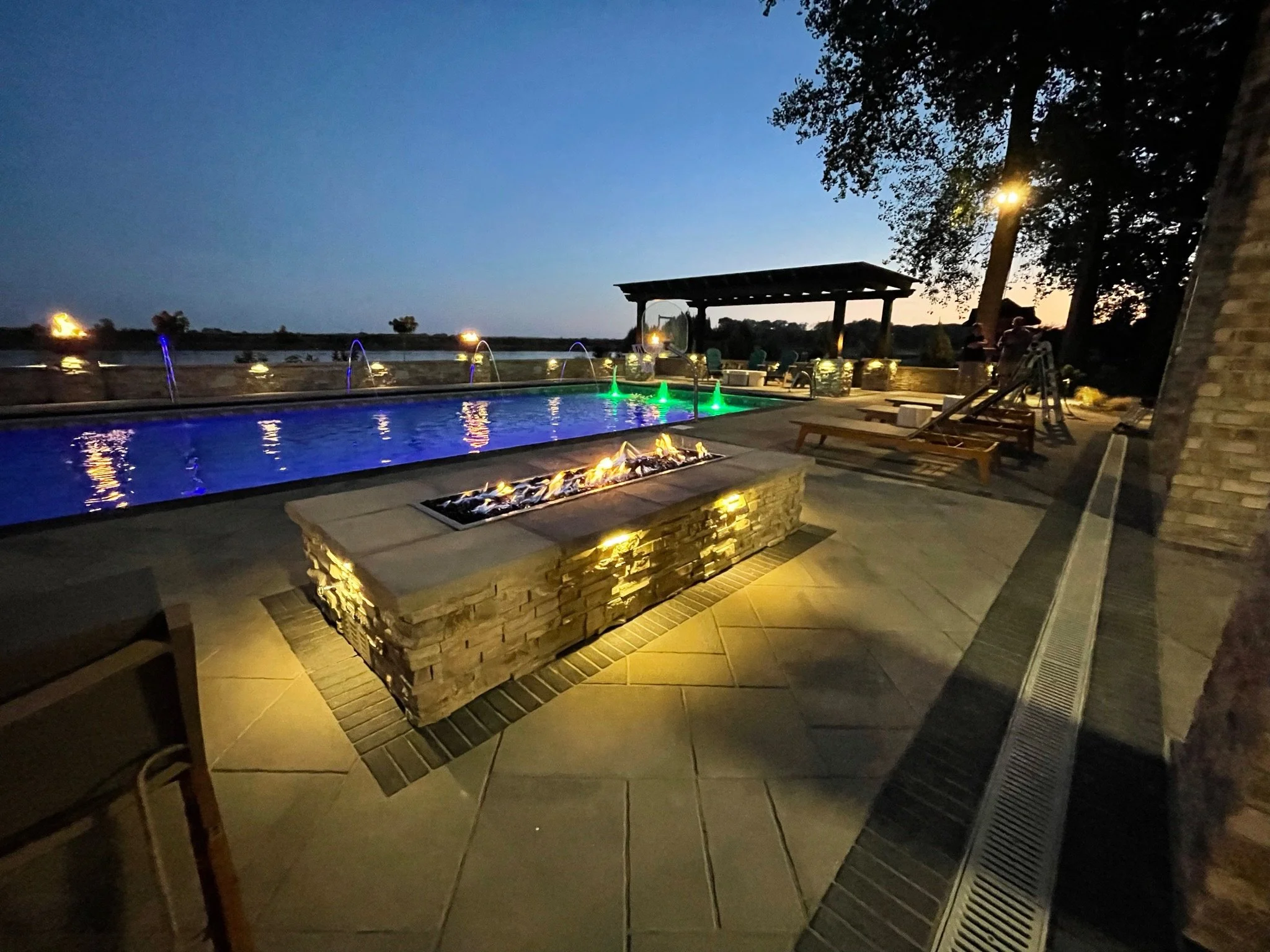 A backyard pool area illuminated at dusk with colorful underwater lights, a fire feature with flames, lounge chairs, a shaded seating area, and a slide in the background.