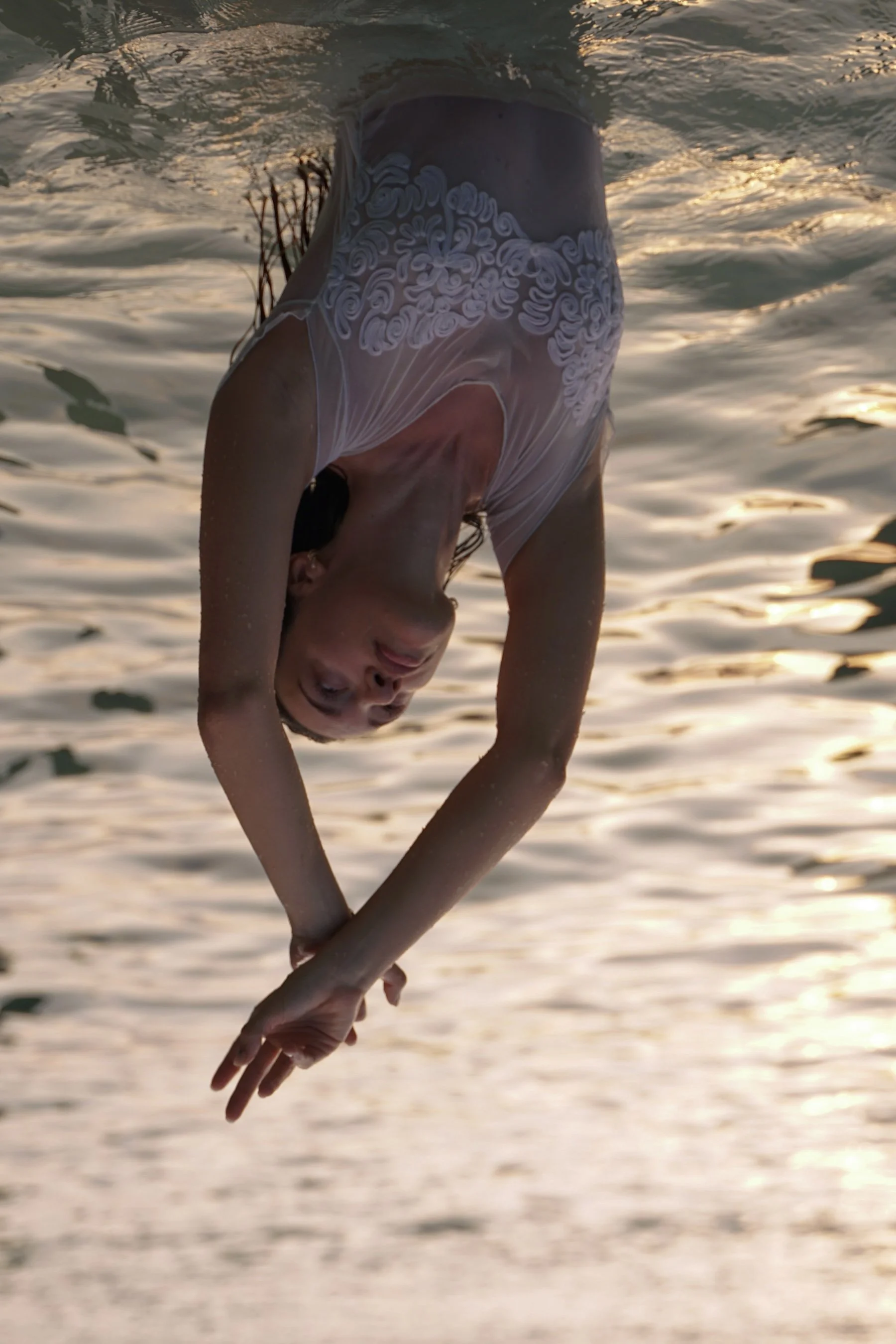 A woman in a sheer, embroidered dress doing a handstand in the water during sunset.