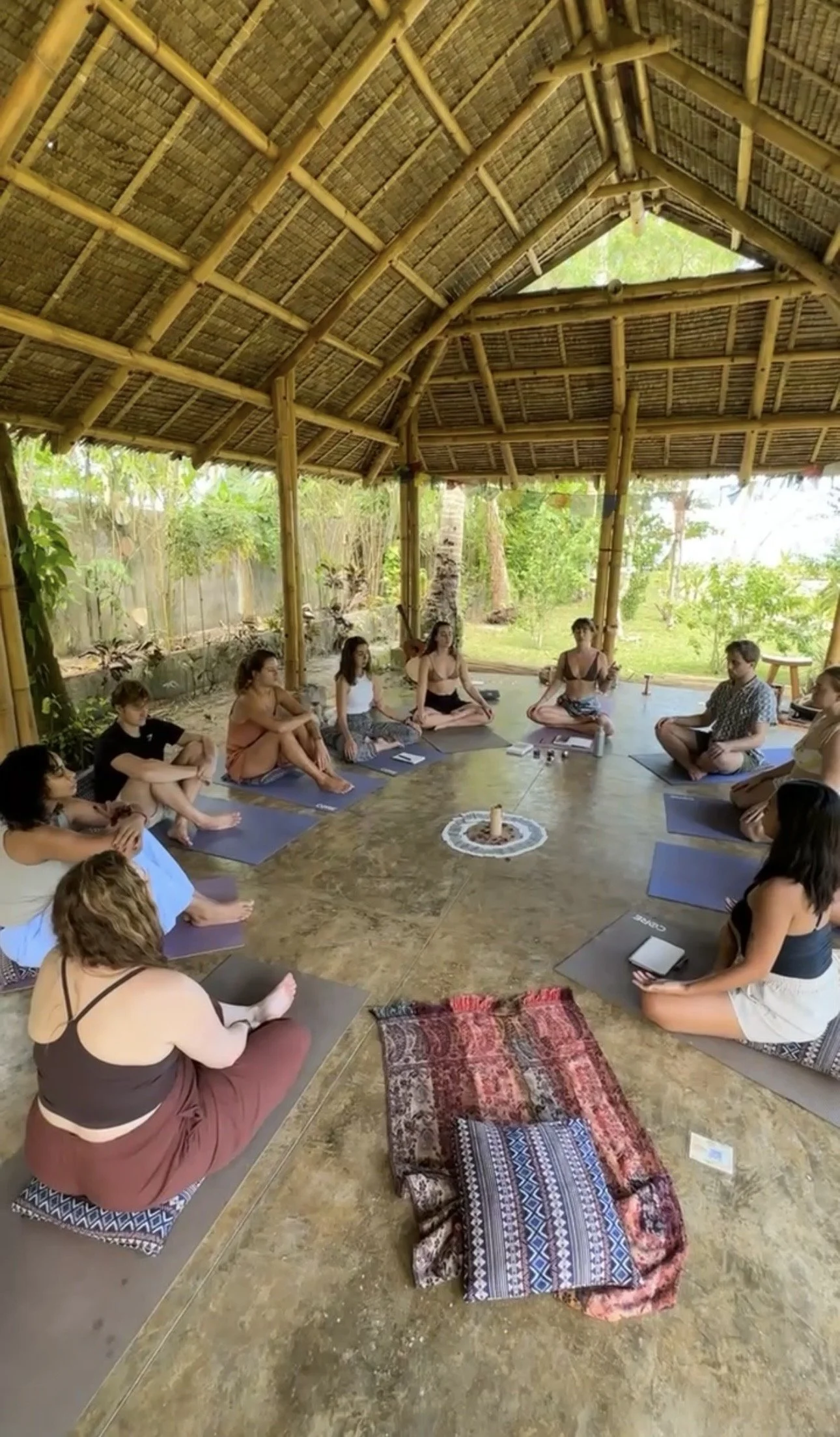 People participating in a group workshop combining breathwork meditation yoga session outdoors under a thatched roof structure, sitting on yoga mats, surrounded by lush green plants and trees.
