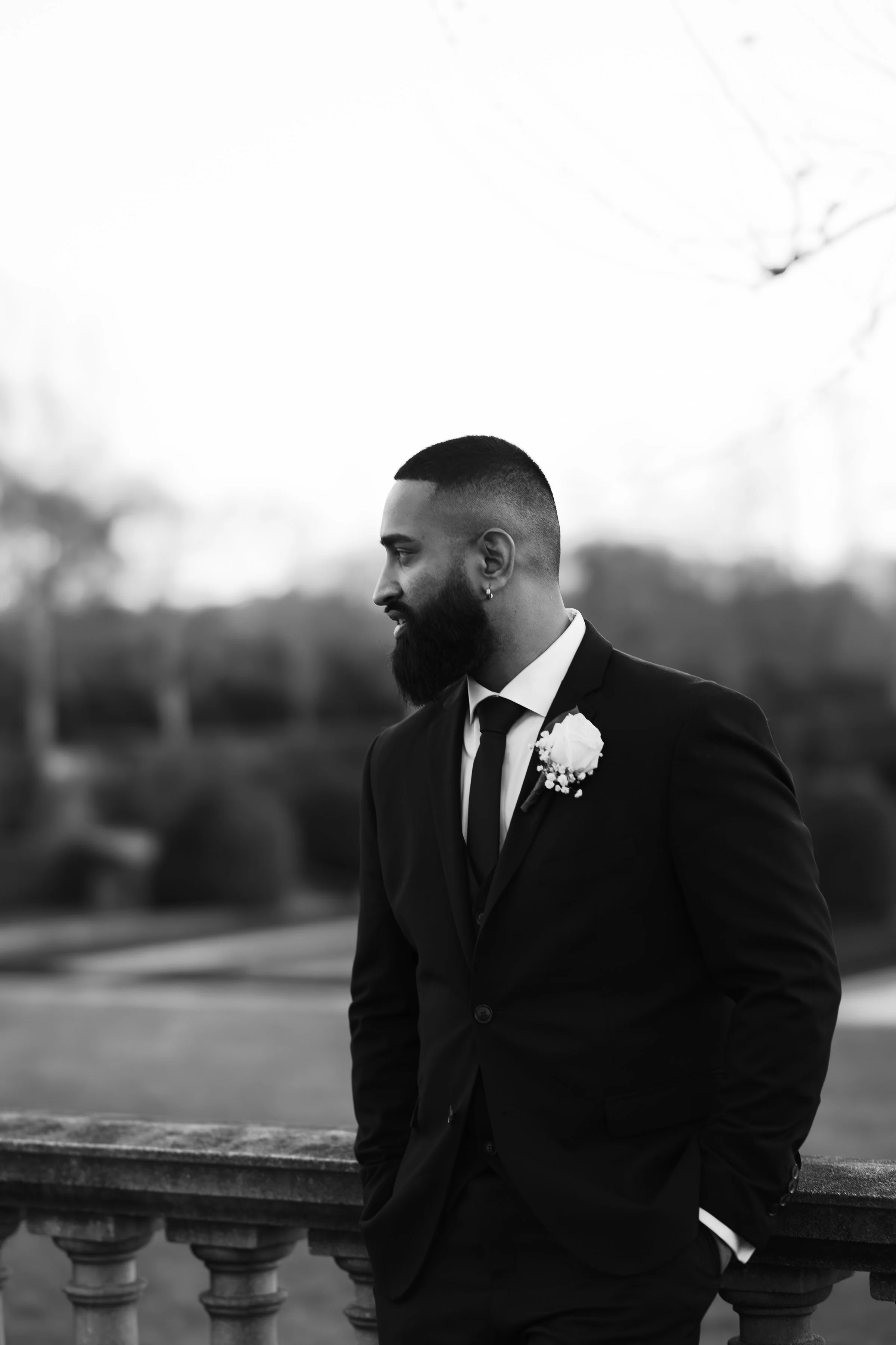 A black and white photo of a man in a suit with a boutonniere, standing outdoors beside a railing, looking to the left.