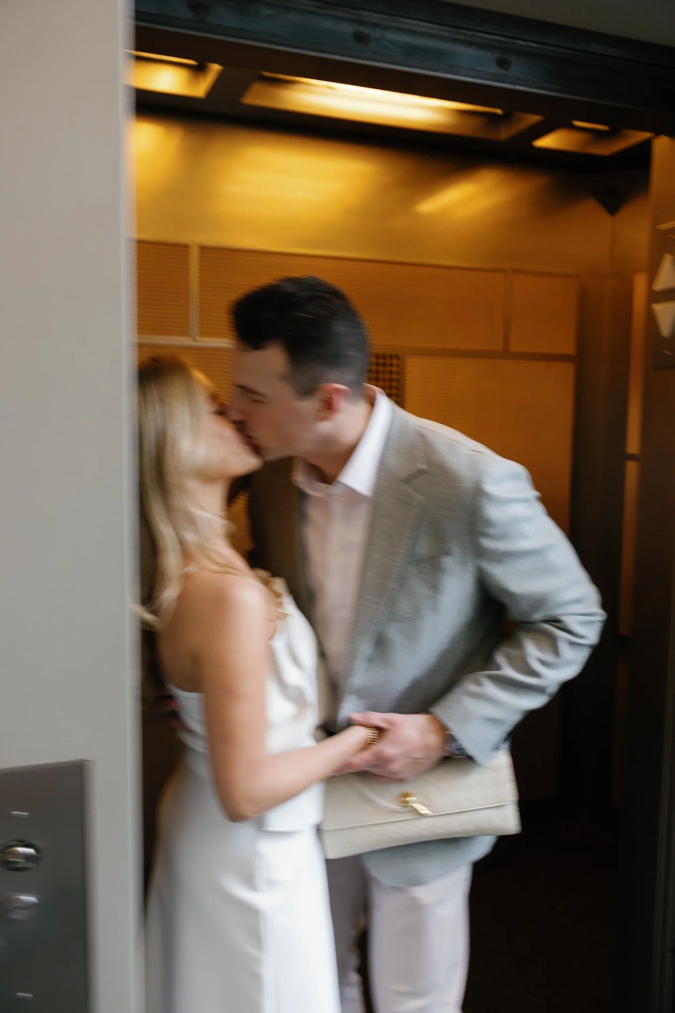 A man and woman sharing a kiss in an elevator, woman holding a clutch purse.
