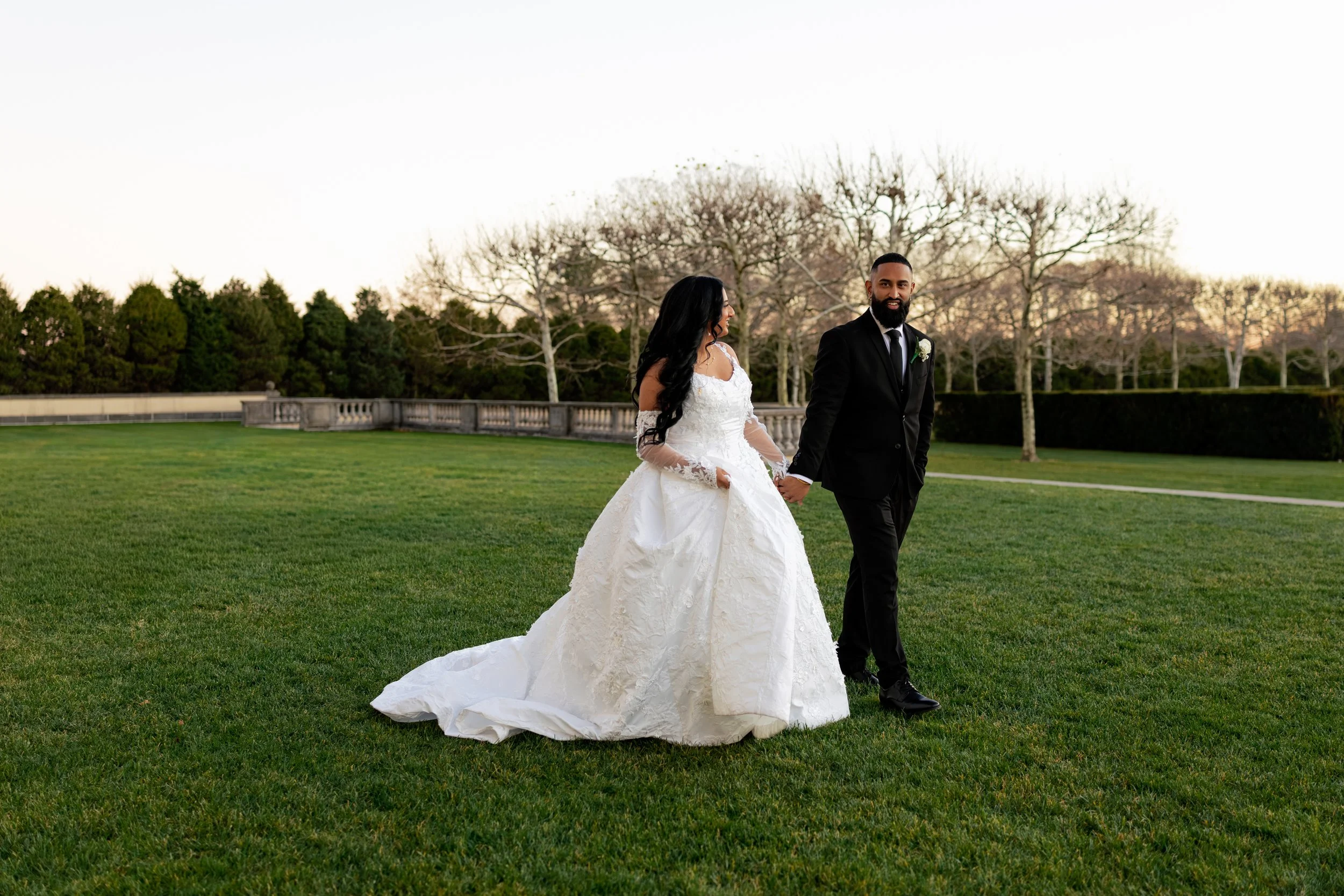 Bride and groom holding hands, walking on grass in an outdoor setting, with bare trees and a stone railing in the background, during sunset.