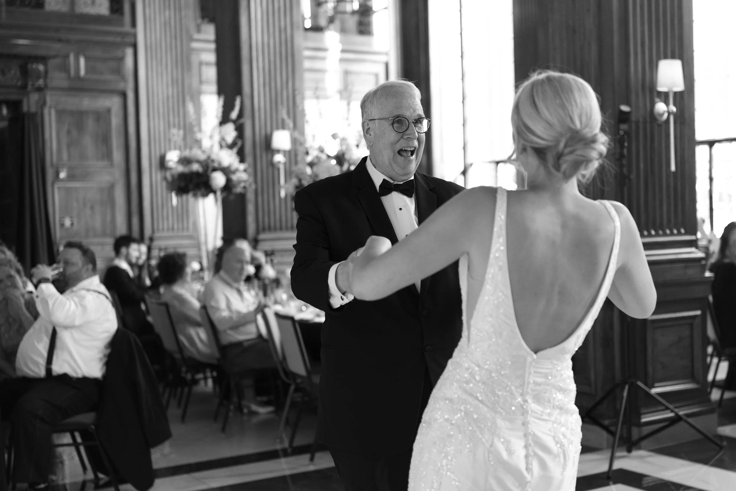 A black-and-white photo of a joyful moment at a wedding reception, showing a man in a tuxedo and glasses dancing with a woman in a white wedding gown inside a decorated hall.