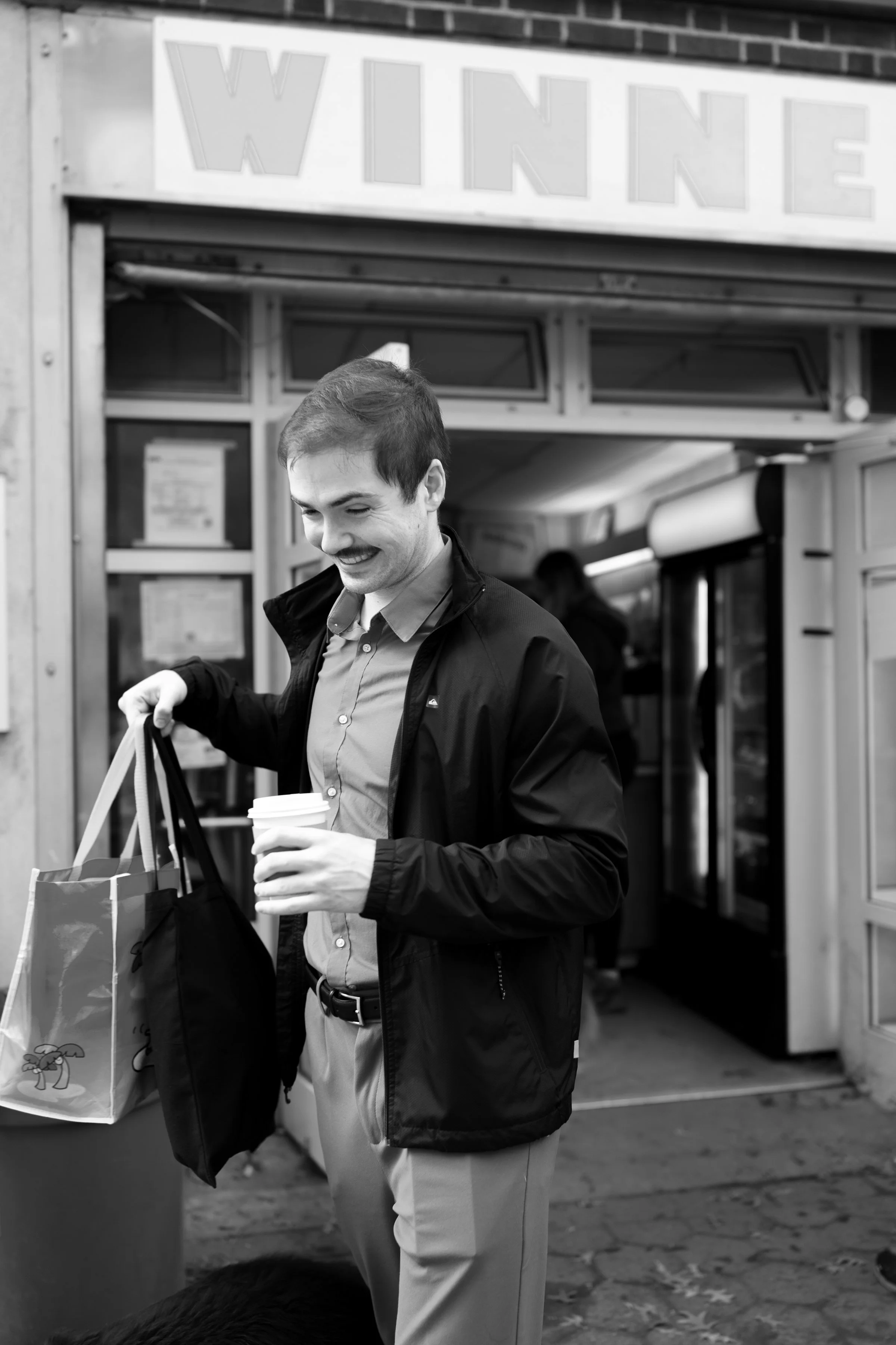 A smiling man holding a coffee cup and shopping bags standing outside a store with a sign reading 'WINNE' in black and white.