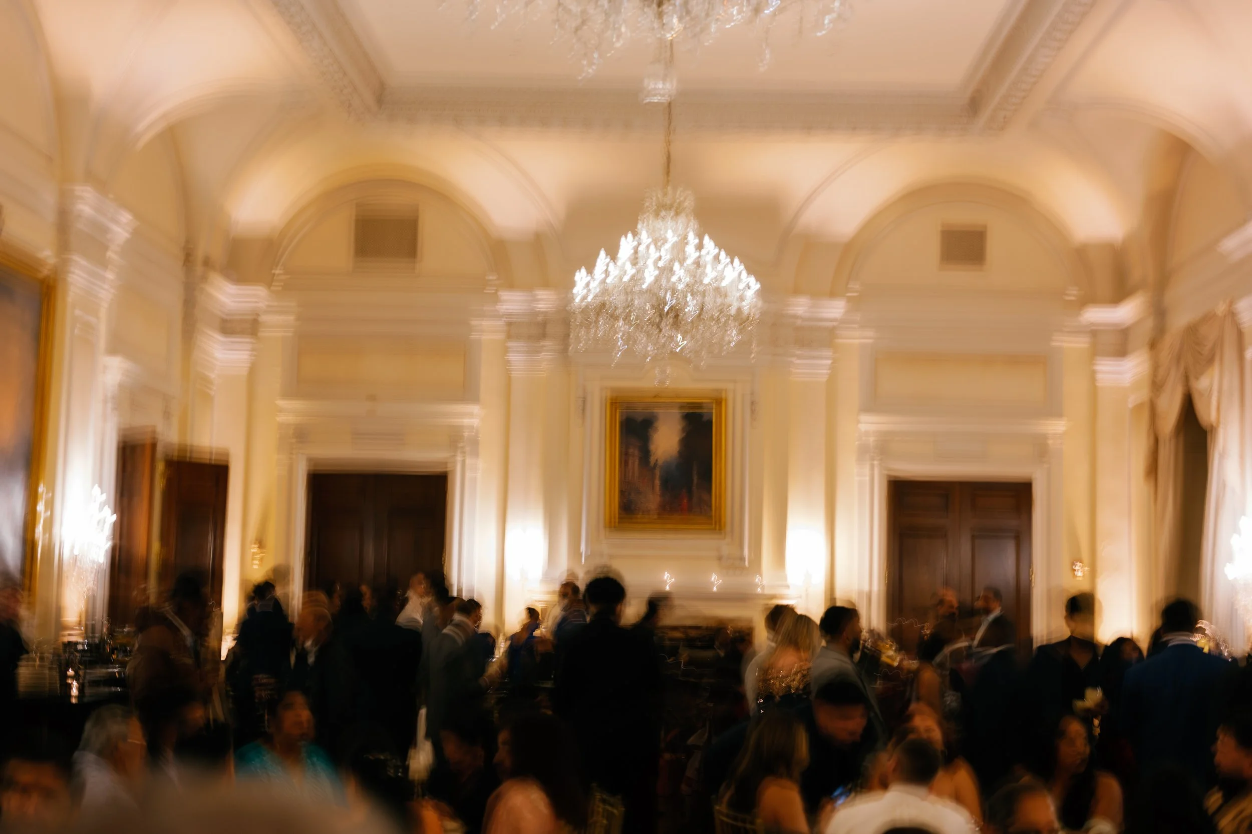 A blurred photograph of an elegant, well-lit ballroom filled with people. The room features tall, arched ceilings, a large chandelier hanging in the center, and ornate decor, including a framed painting above a fireplace.