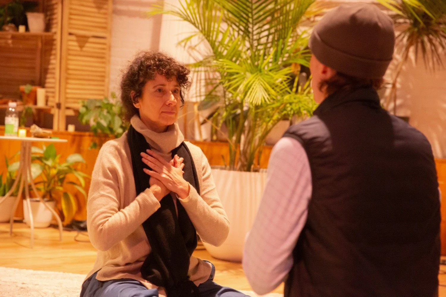 Two people sitting on the floor in a warm, cozy room with wooden shelves, large plants, and soft lighting, engaged in a heartfelt conversation. The woman faces the camera with her hands over her chest, appearing emotional. The man has his back to the