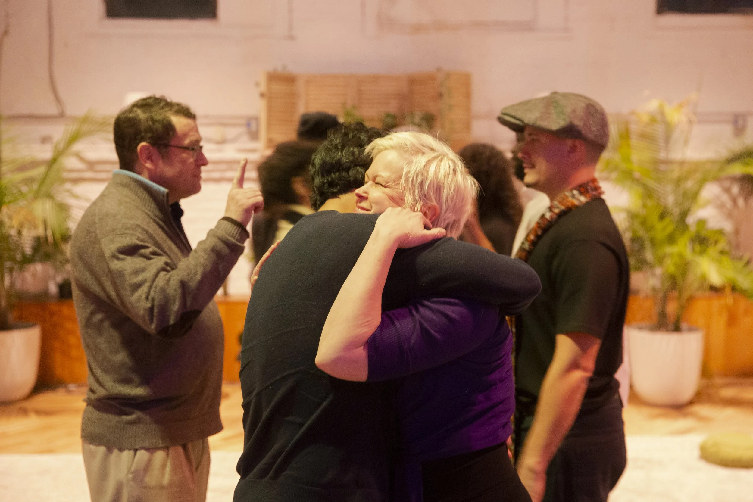 Two women hugging and smiling, surrounded by other people in a warmly lit indoor setting.