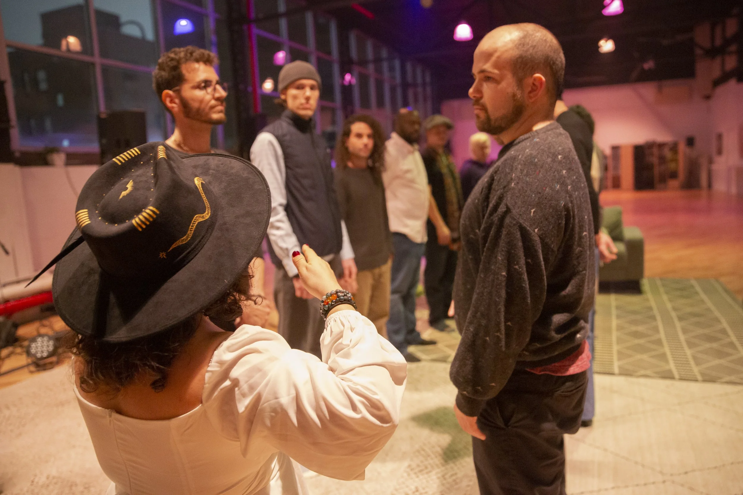 Group of people in a room with large windows and purple lighting, standing in a line and facing each other, with a woman in a black hat with a snake design and a cream-colored dress gesturing toward a man with a beard in a dark sweater, possibly duri