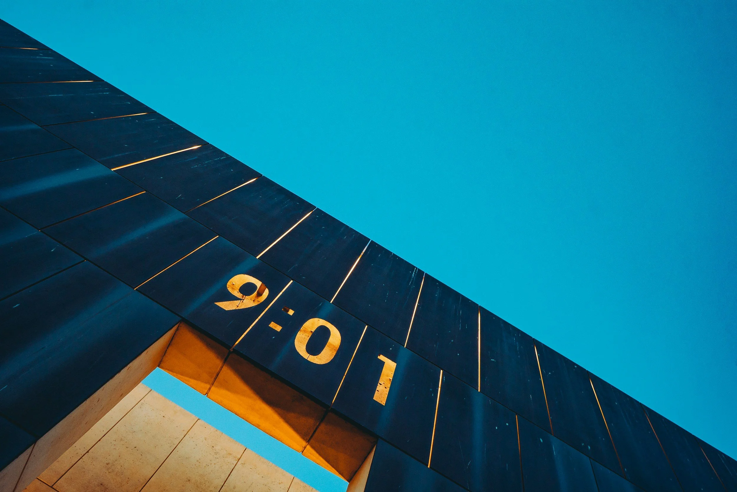 Architectural detail showing a metal panel structure with the time "9:01" cut out, against a blue sky.