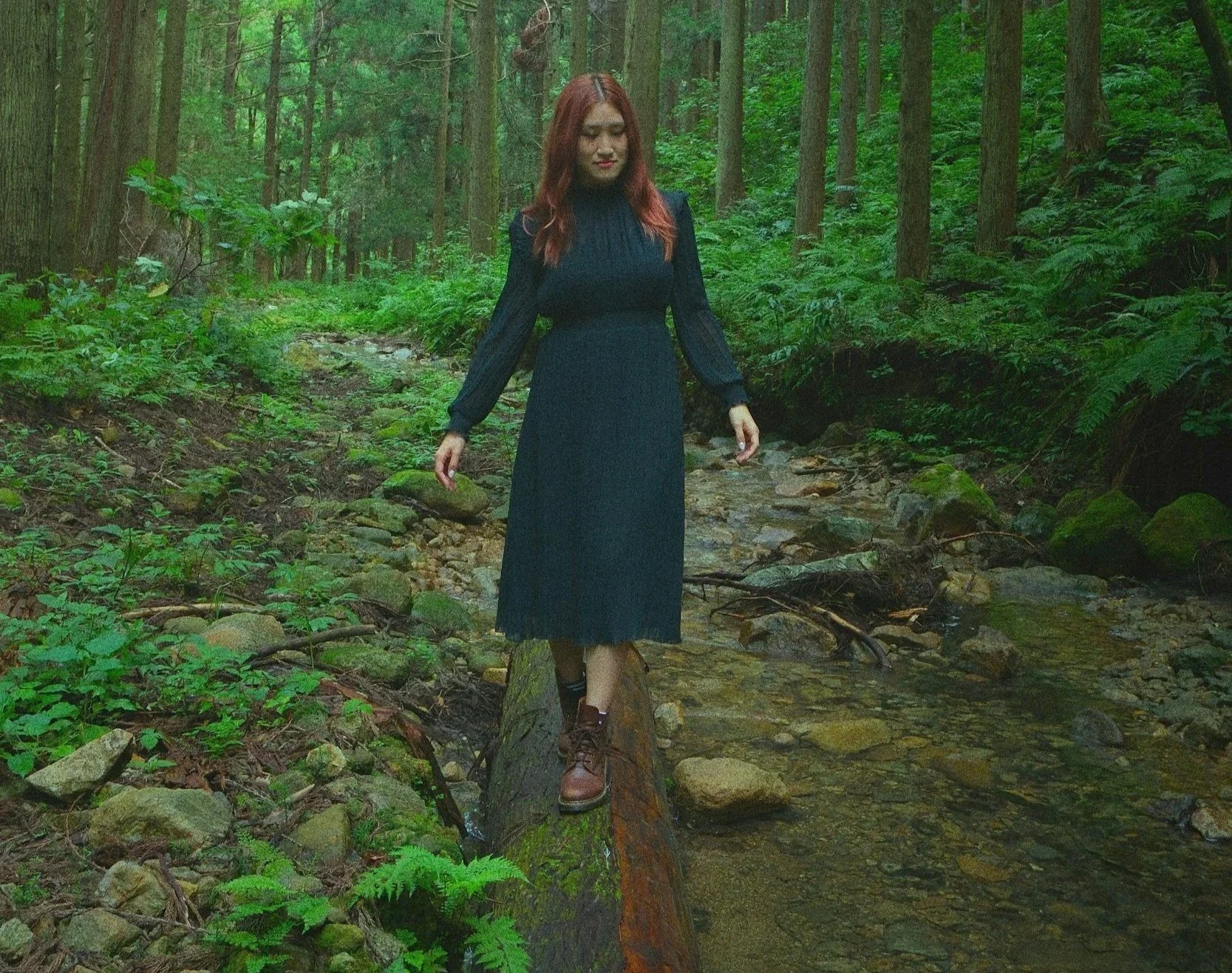A person in a dark dress and boots balances on a log on a stream in the forest, symbolizing overcoming enmeshment