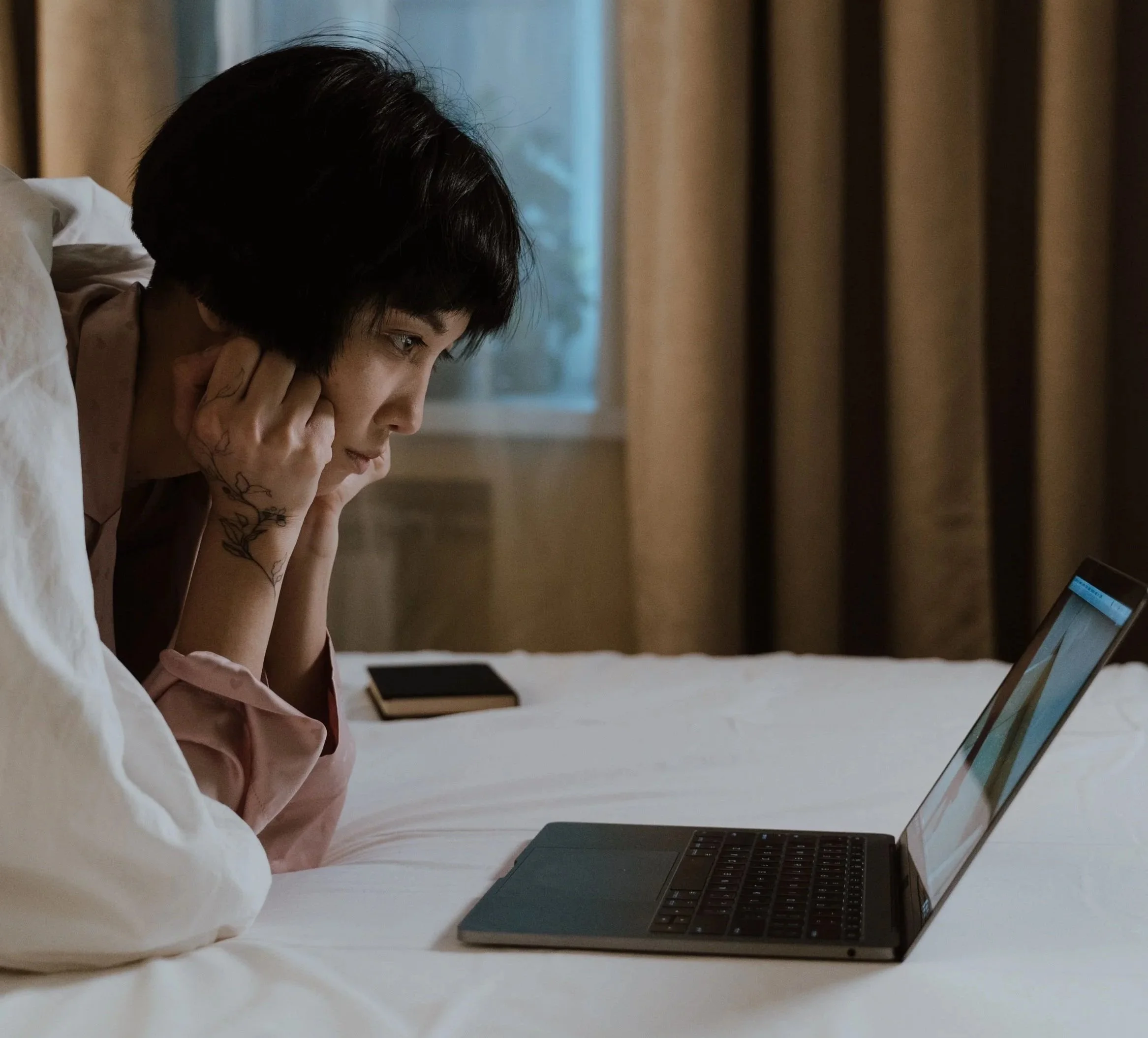 A person with short black hair and hangs is under the covers in bed, chin prompt up on their hands. They are looking at a laptop screen.