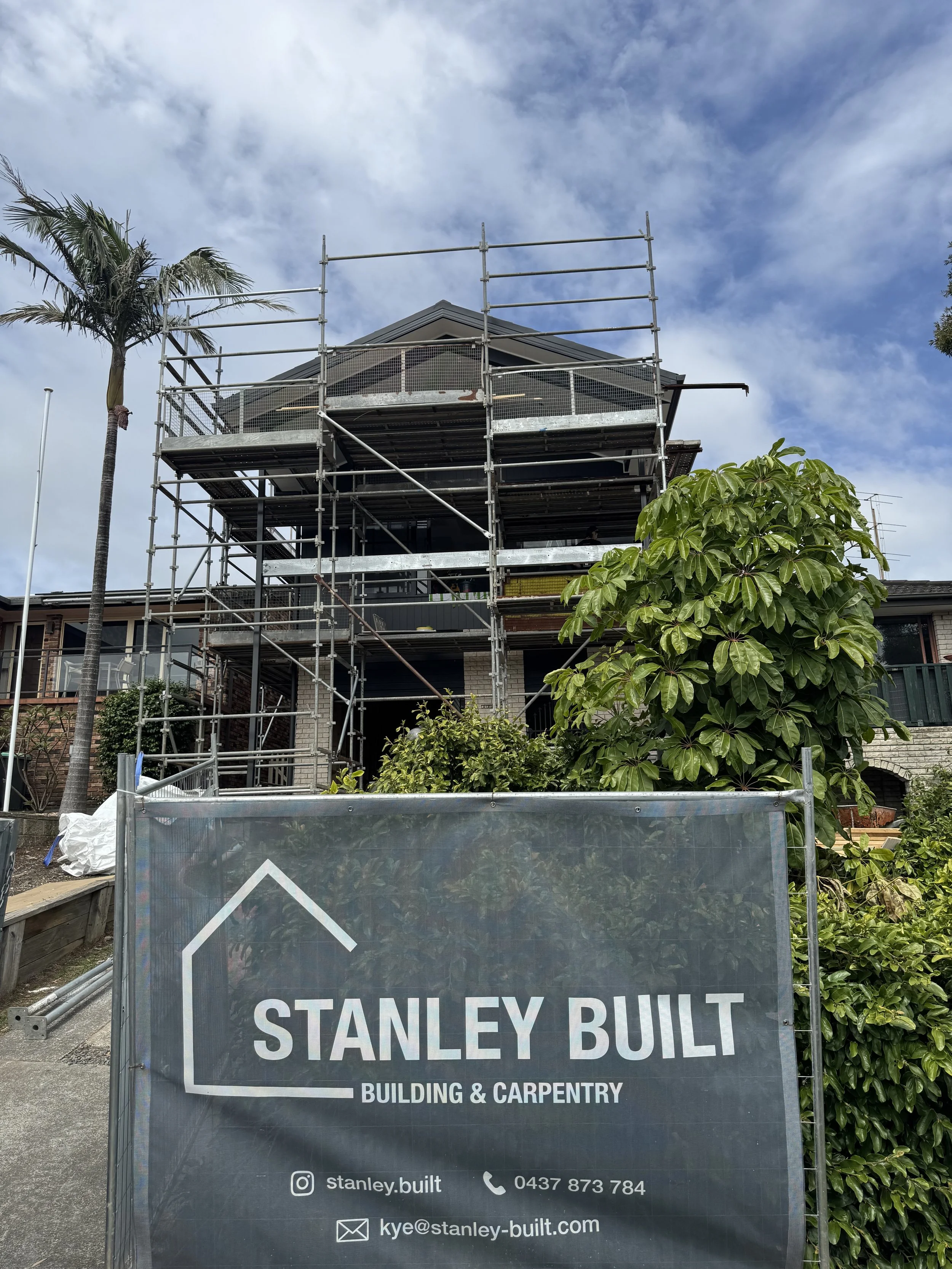A building under construction with scaffolding around it. In the foreground, there is a sign for Stanley Built, a building and carpentry company. The sign displays contact information and social media handle. There are trees and partly cloudy skies in the background.