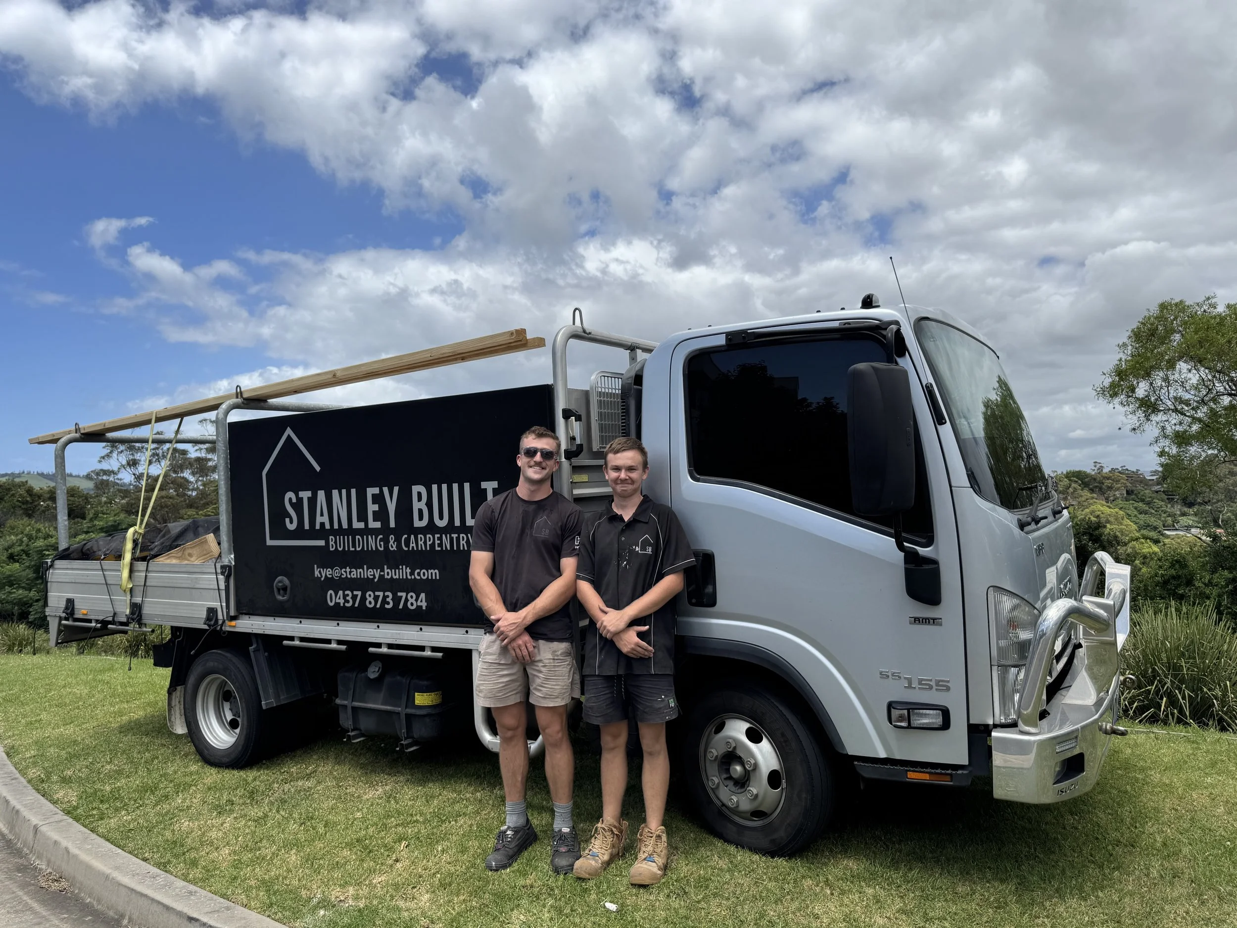 Two young men standing in front of a white work truck with a black sign reading "Stanley Built Building & Carpentry". The truck bed has wooden planks and equipment. The background includes green trees, a grassy area, and a partly cloudy sky.