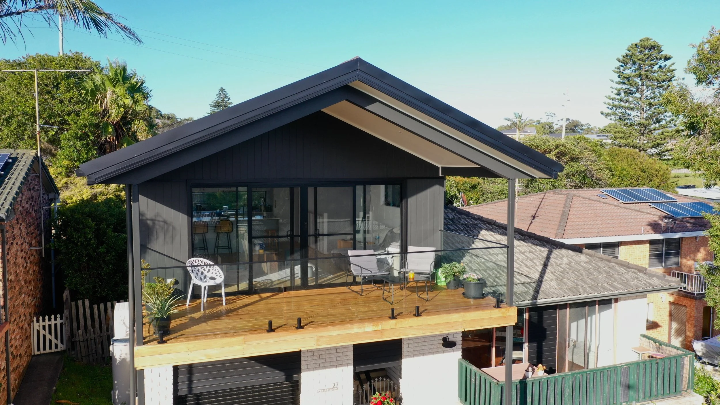 Modern house with a rooftop deck featuring outdoor furniture, potted plants, and glass railing, set against a backdrop of trees and neighboring rooftops with solar panels.