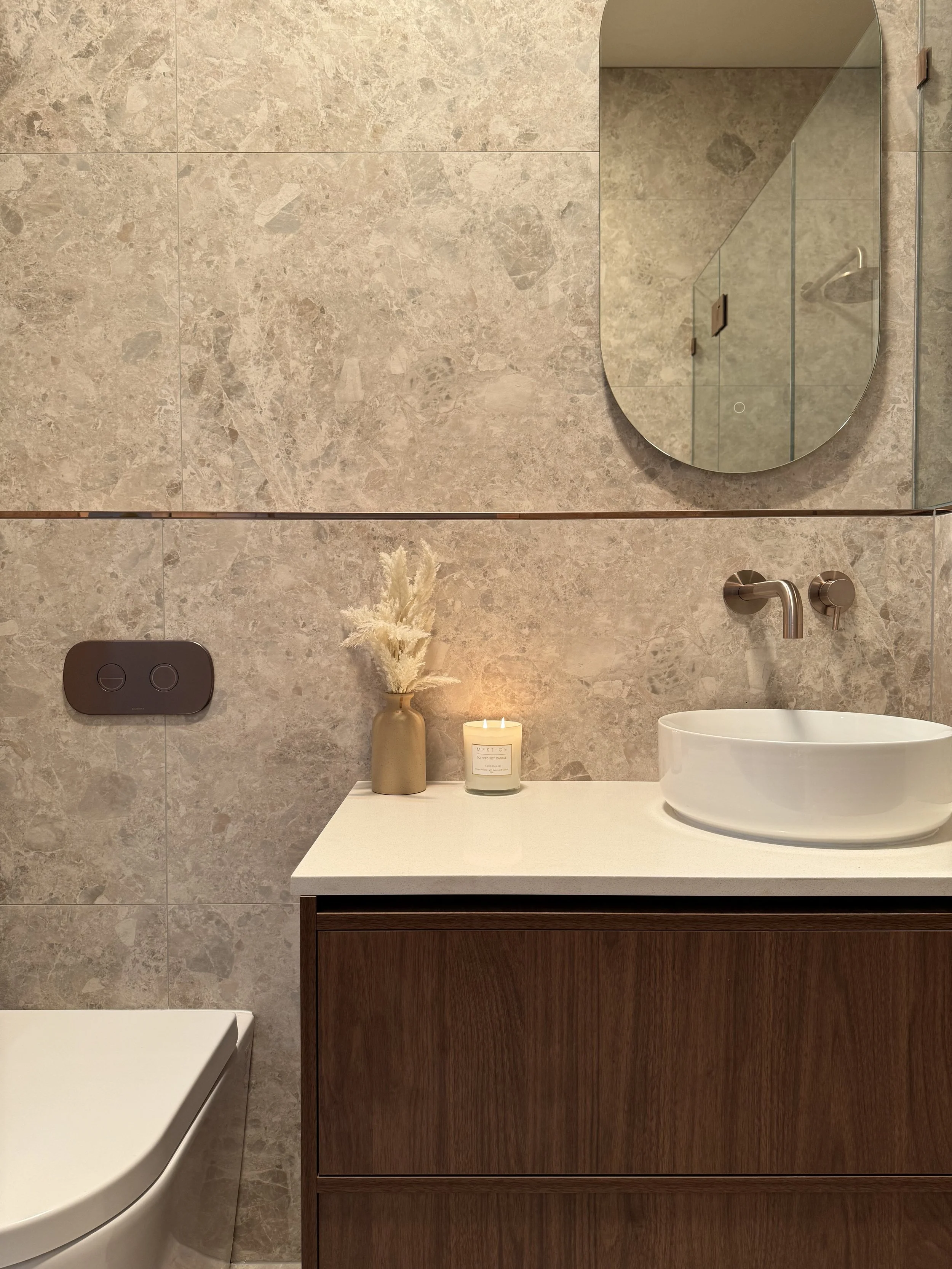 Modern bathroom with beige marble tiled wall, a wooden vanity with a white countertop, a round vessel sink, a mirror, a candle, a vase with pampas grass, and a wall-mounted faucet.