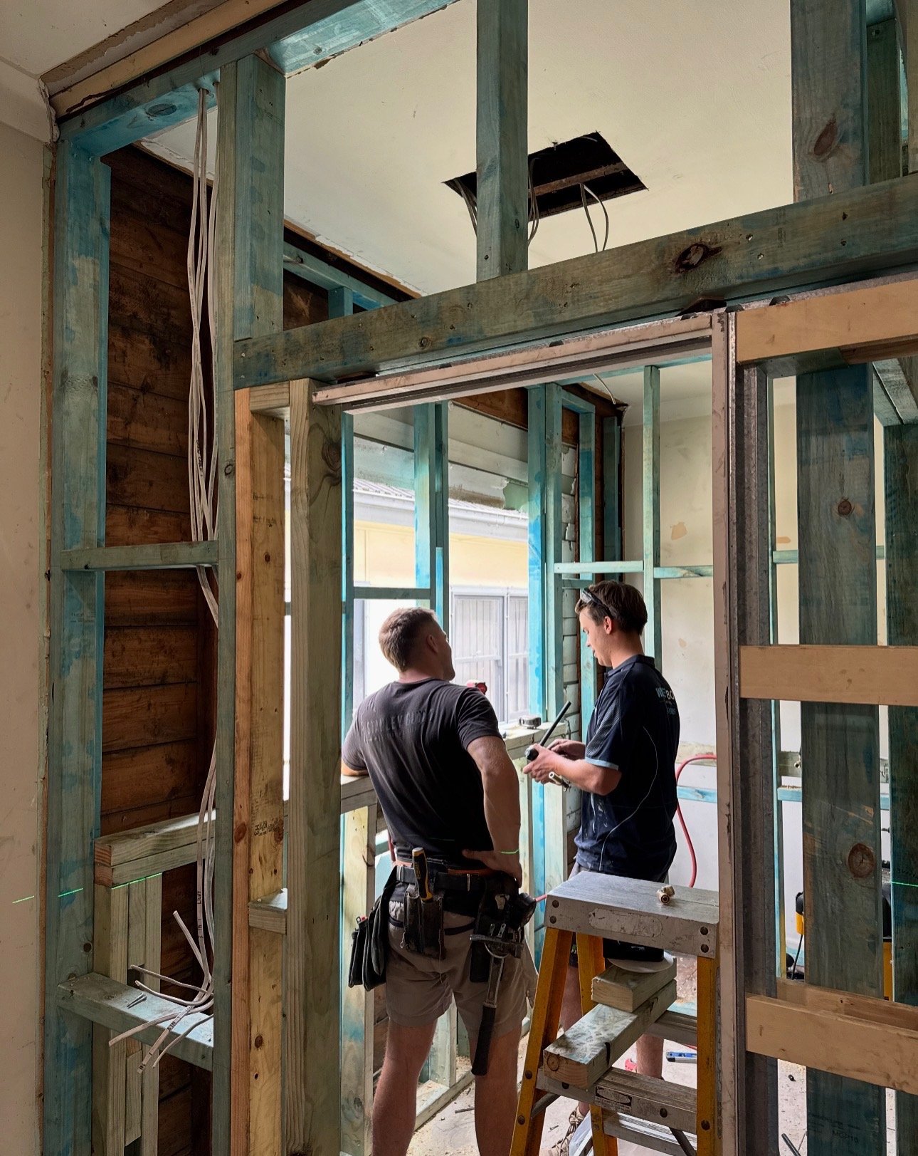 Two construction workers inside a house under renovation, surrounded by wooden framing and construction tools, having a discussion.