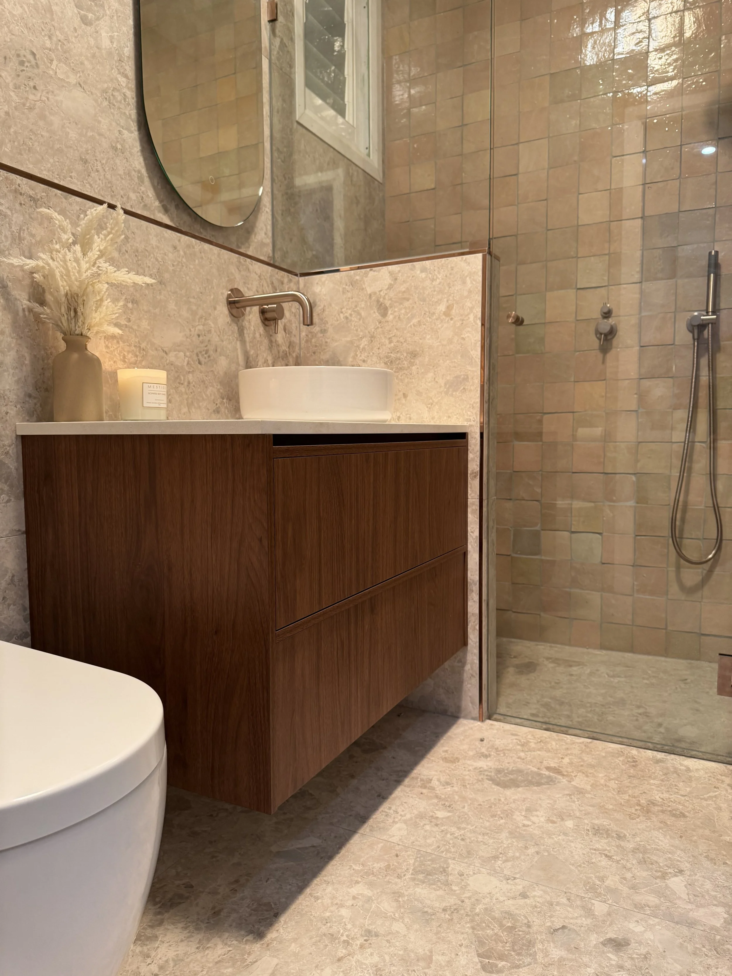 Modern bathroom with beige tiled flooring and walls, a wooden vanity with a round vessel sink, a large mirror, decorative dried flowers and candle on the vanity, and a walk-in shower with glass walls and beige tiled interior.