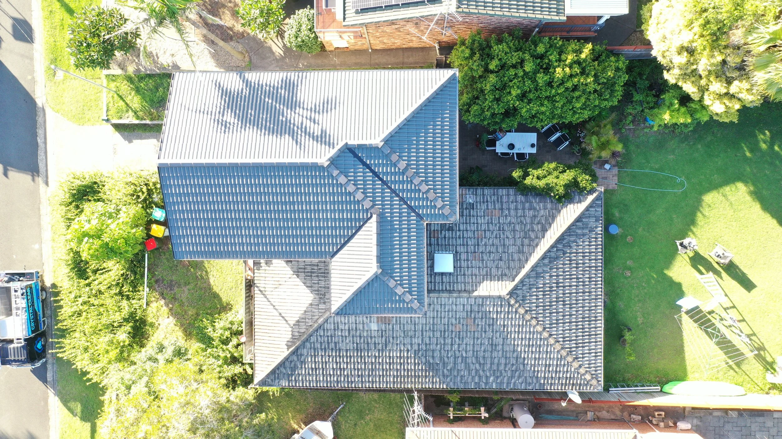 An aerial view of a residential backyard with a house that has a gray metal roof, green trees, a patio with outdoor furniture, and a grassy lawn with two chairs.