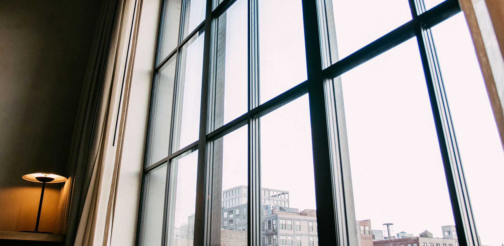 Interior view of a room with large windows looking out onto city buildings, with a lamp on a side table nearby.
