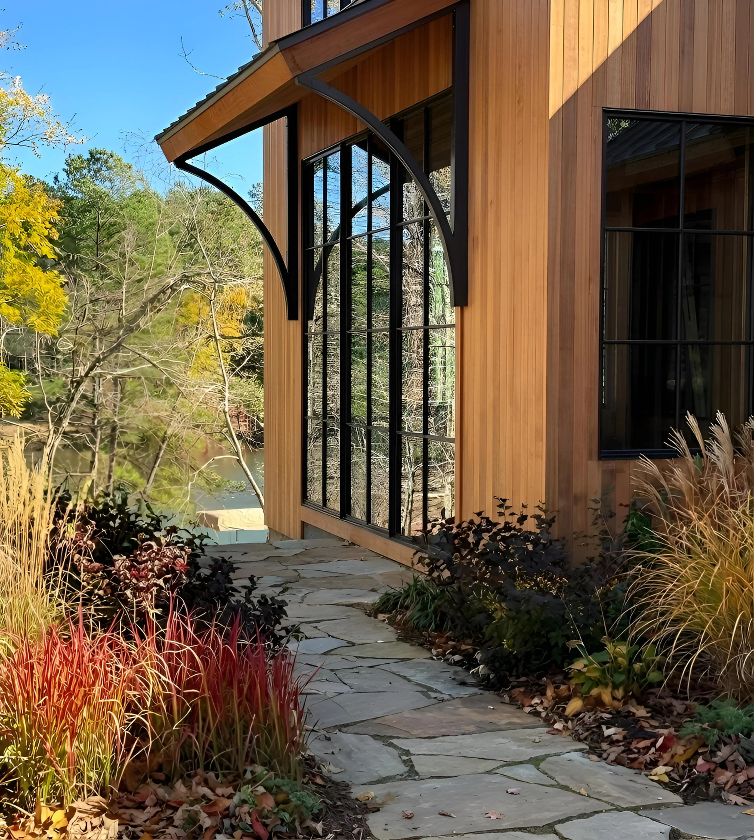 A house with large black-framed windows and light wood siding, surrounded by autumn plants along a stone pathway, with trees and a river in the background under a clear blue sky.