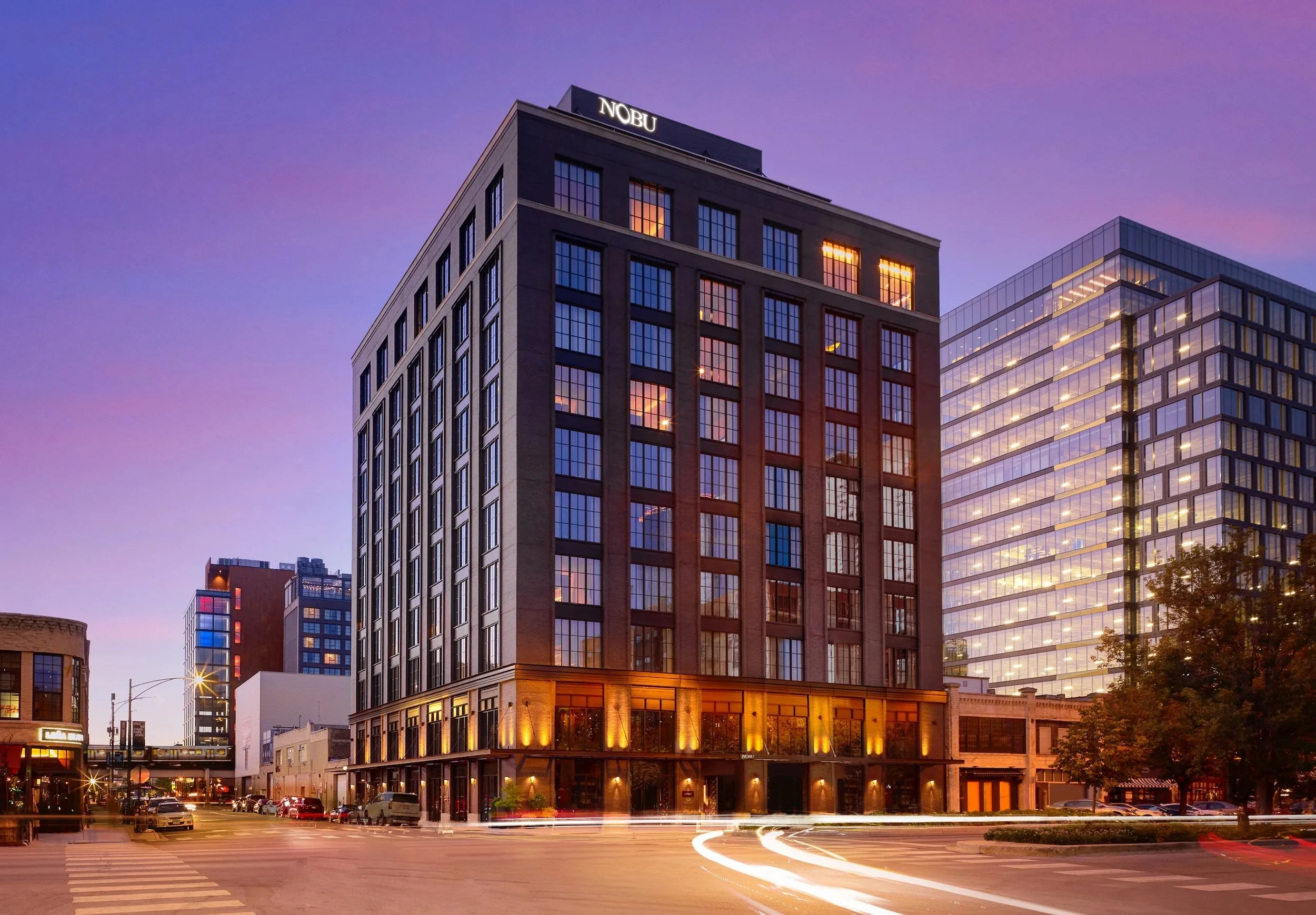 Cityscape at dusk featuring a tall building with illuminated windows and the sign 'NOBU' on top, surrounded by modern glass buildings, with traffic streaks on the street in the foreground.