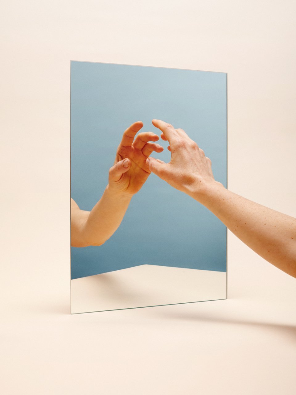 Two hands reach for each other reflected in a mirror, with a light blue background.