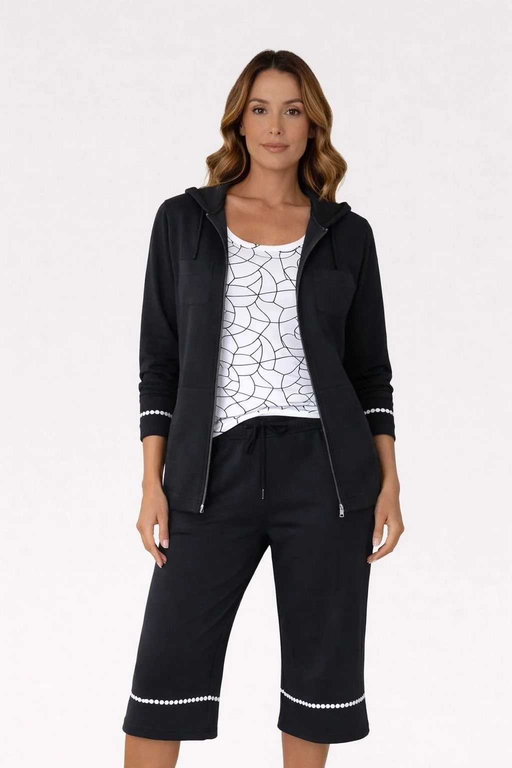 A woman wearing a black zip-up hoodie, white patterned t-shirt, and black cropped pants standing against a plain white background.