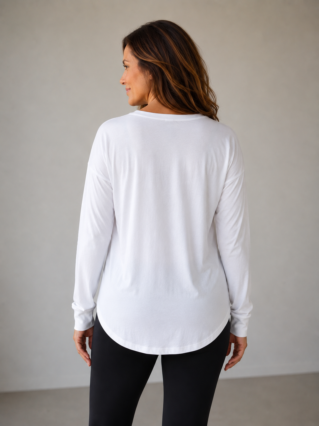 Woman in a white long-sleeve shirt, seen from the back, with brown wavy hair, standing against a plain wall.