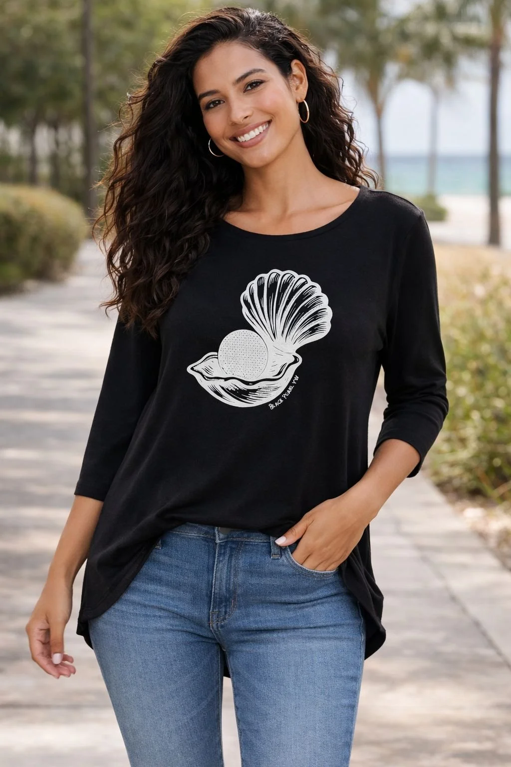 Woman with curly hair smiling outdoors, wearing a black long-sleeve t-shirt with a graphic of a shell and a round object inside, and blue jeans
