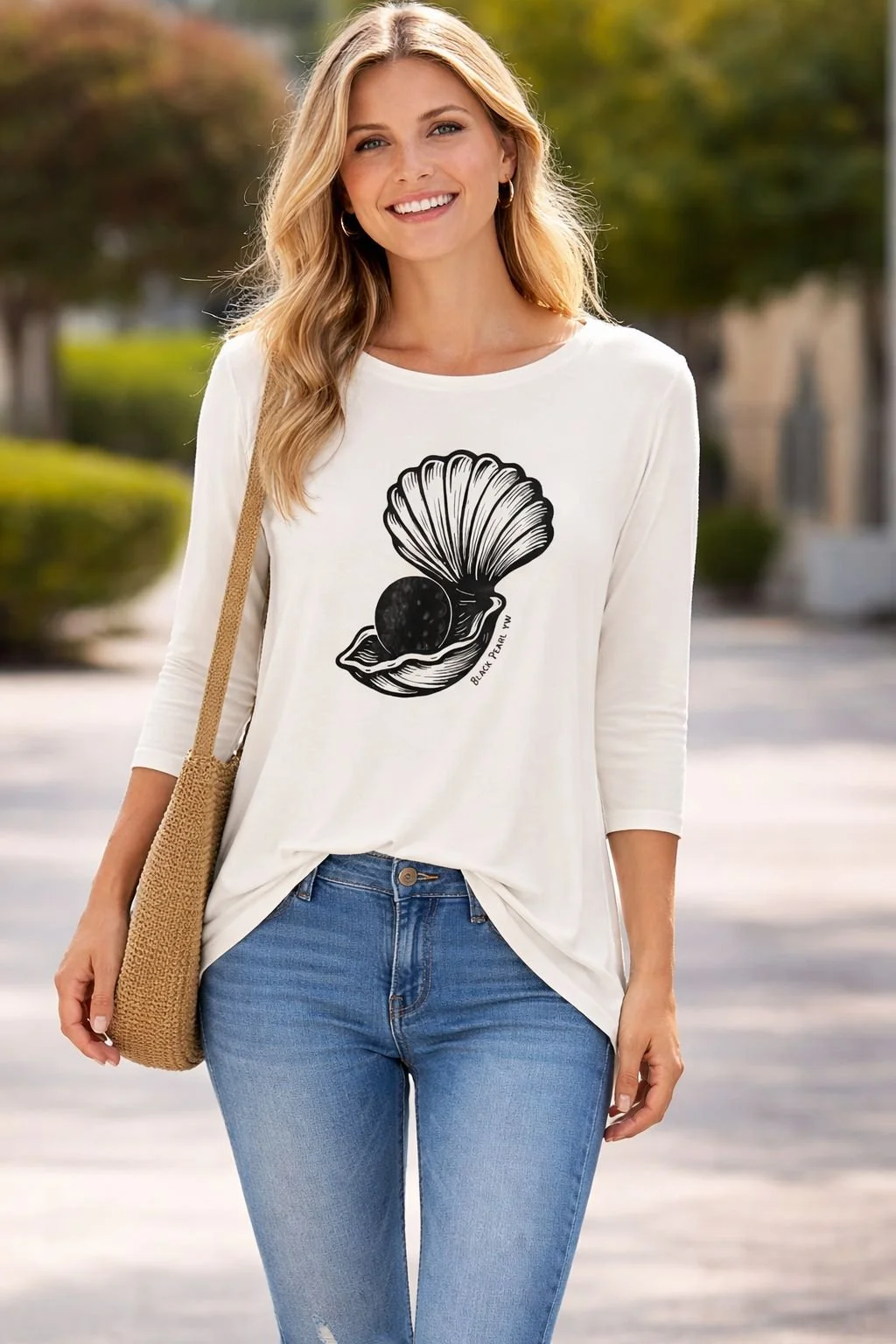 A woman smiling outdoors wearing a white long-sleeve T-shirt with a shell and pearl design and blue jeans.