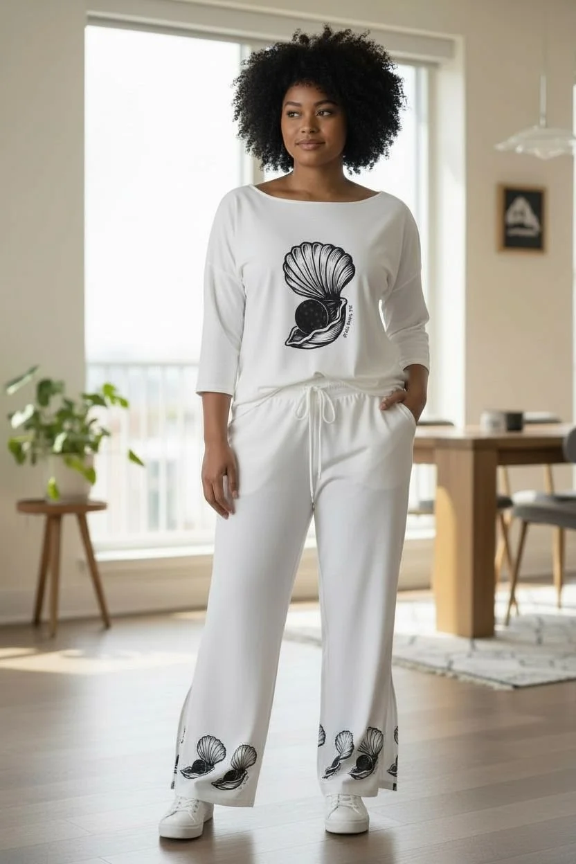 A woman with curly black hair standing in a bright, modern living room, wearing a white matching pajamas set with black shell and pearl shell designs.