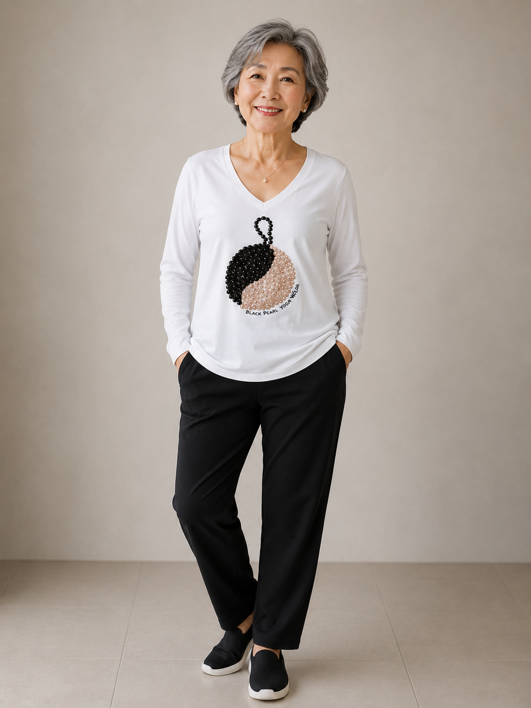 Smiling elderly Asian woman with short gray hair wearing a white long-sleeve shirt with a beaded house design and the words 'Black Pearl Yoga & Wellness', black pants, and black slip-on shoes, standing against a plain beige background.