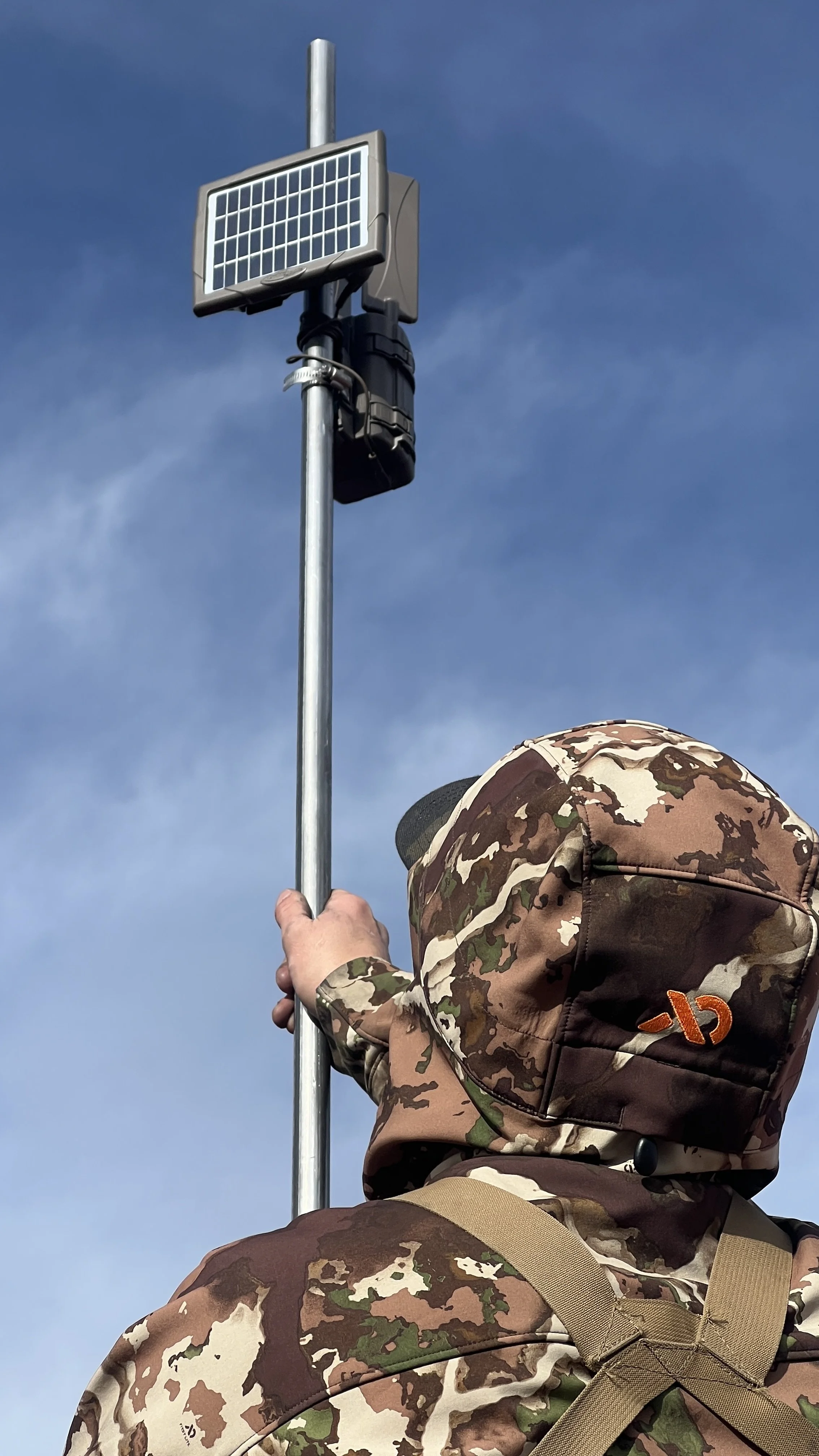 Person in camouflage gear attaching a solar-powered radar or camera device to a pole against a cloudy sky