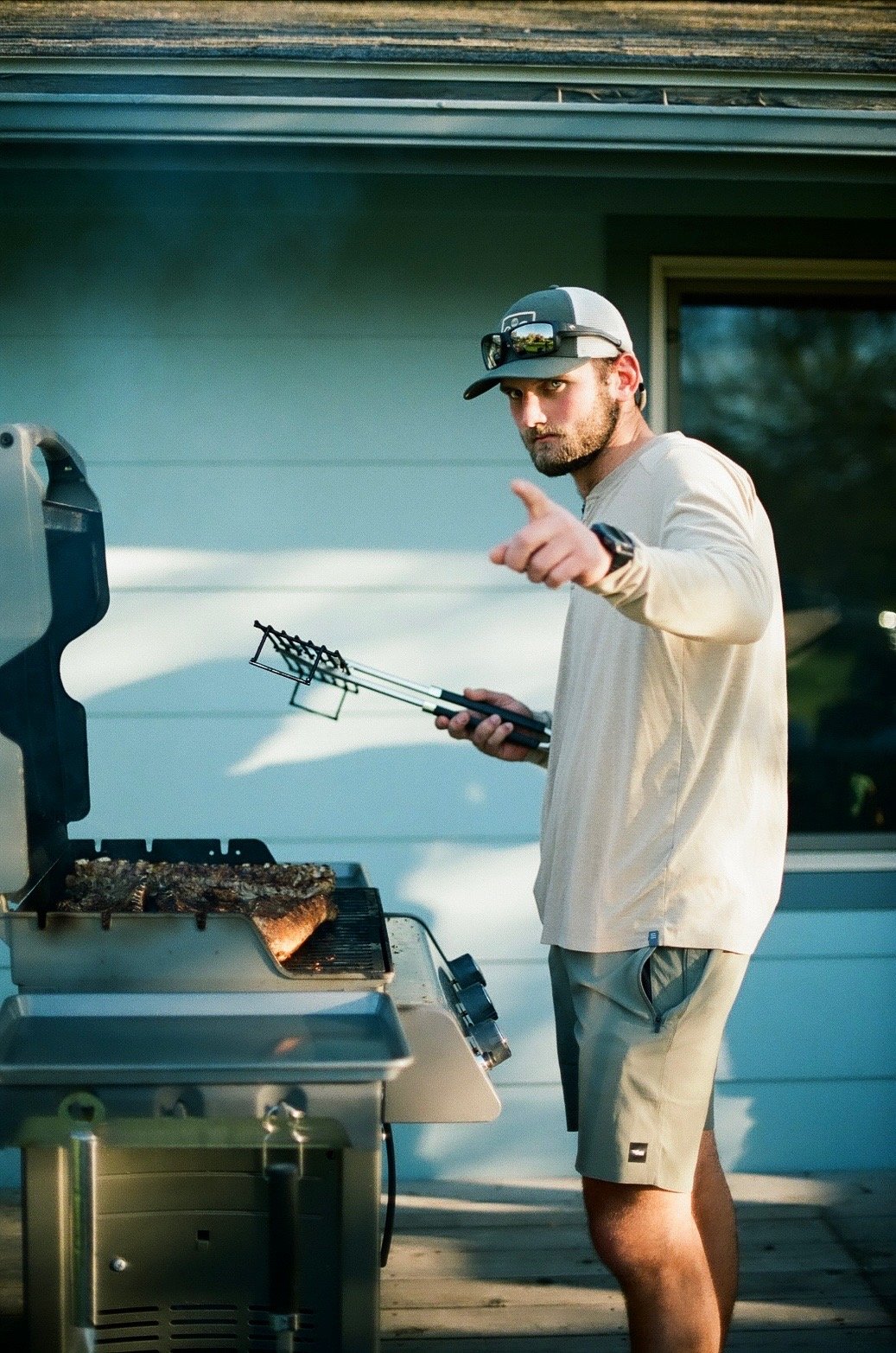 A man grilling meat on a barbecue grill outdoors, pointing at the camera.