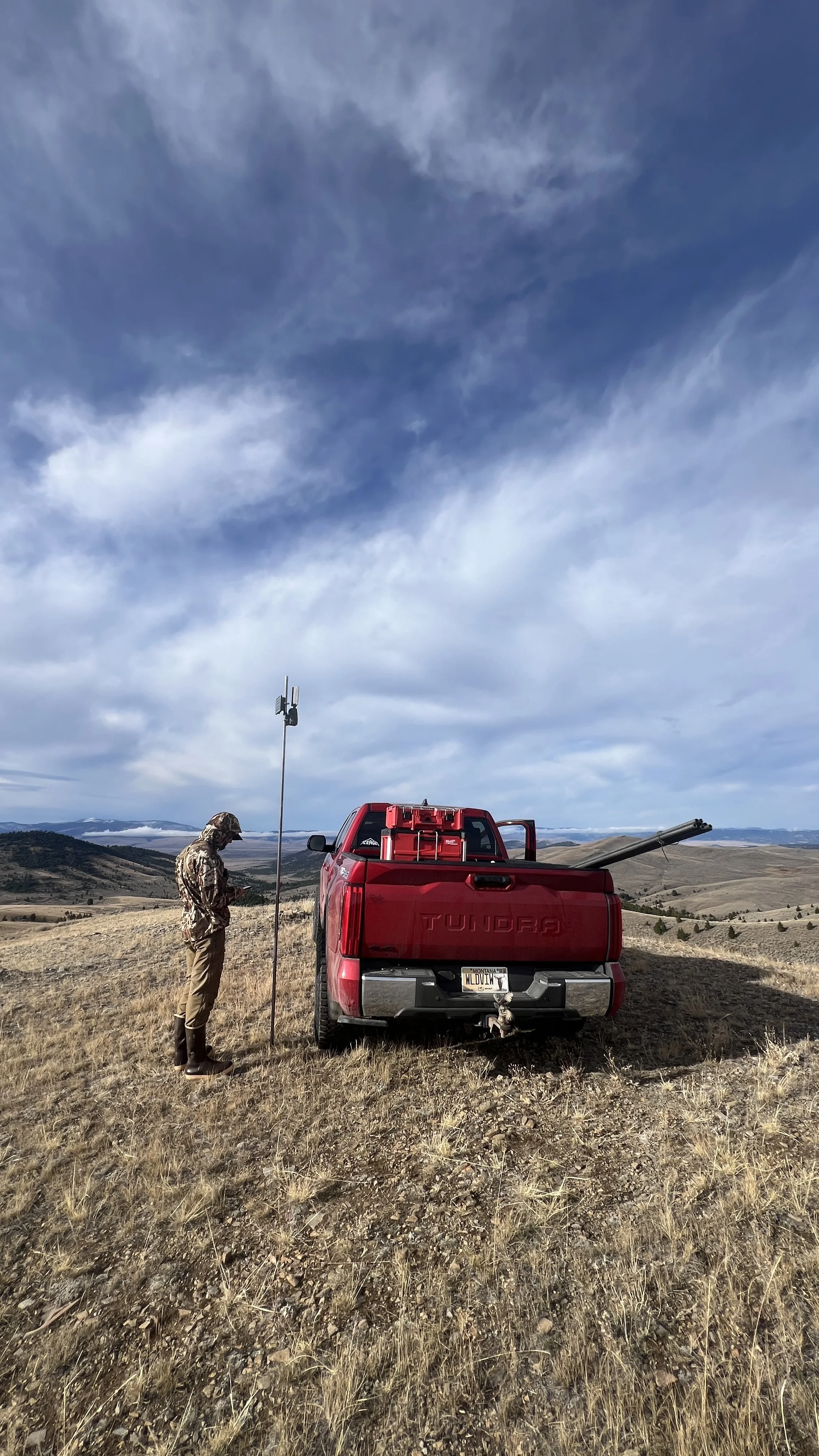 A person in camouflage clothing standing next to a red Toyota Tundra pickup truck on a grassy, hilly landscape under a partly cloudy sky. There is a tall antenna or weather station next to the person.
