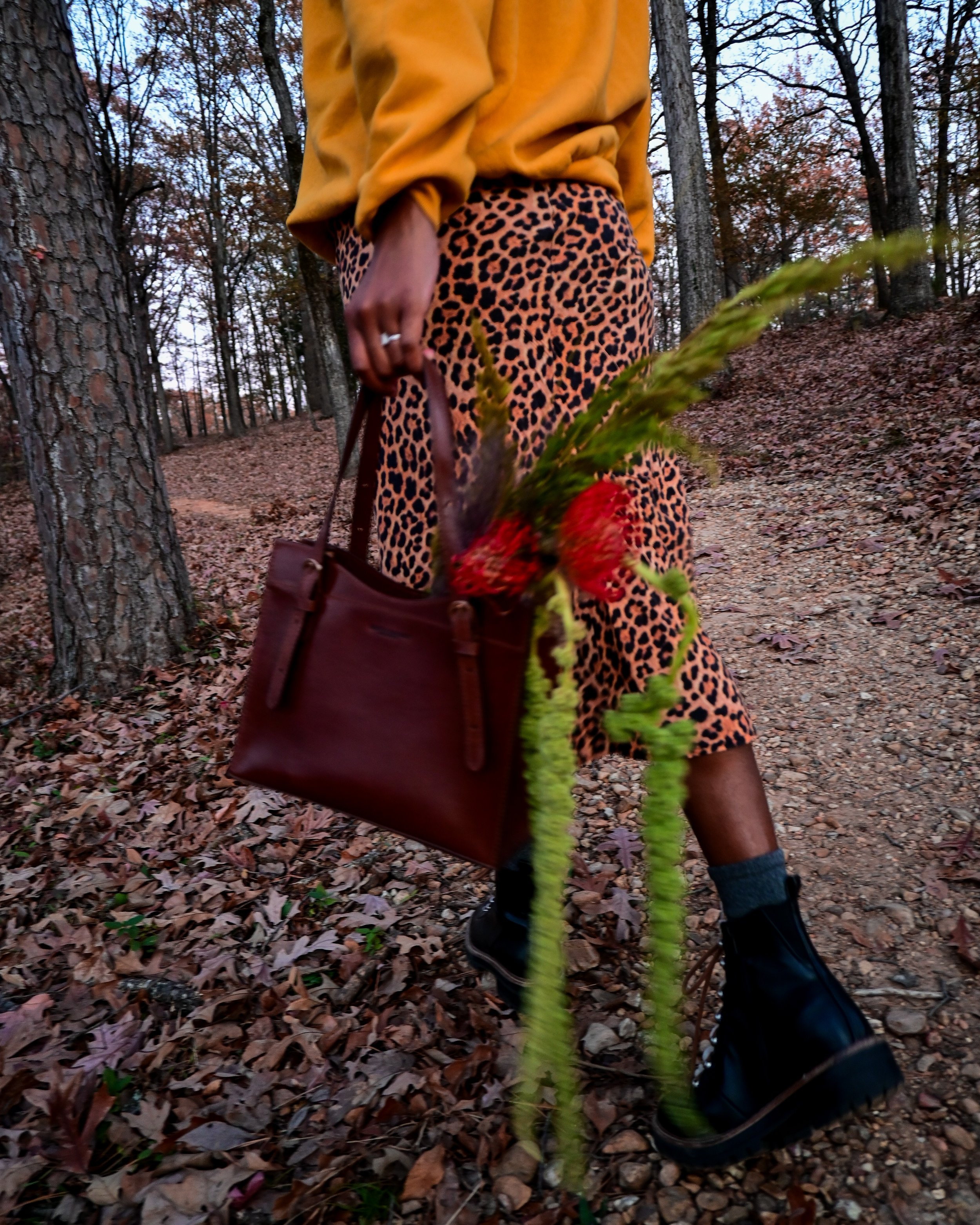 Person walking in a wooded area during autumn, carrying a tote bag with red flowers, wearing a yellow top, leopard print skirt, and black boots.