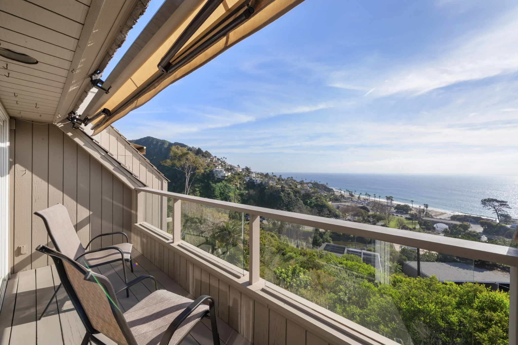 A balcony with three chairs overlooking a scenic view of the ocean, hillside houses, and lush green trees under a partly cloudy sky.