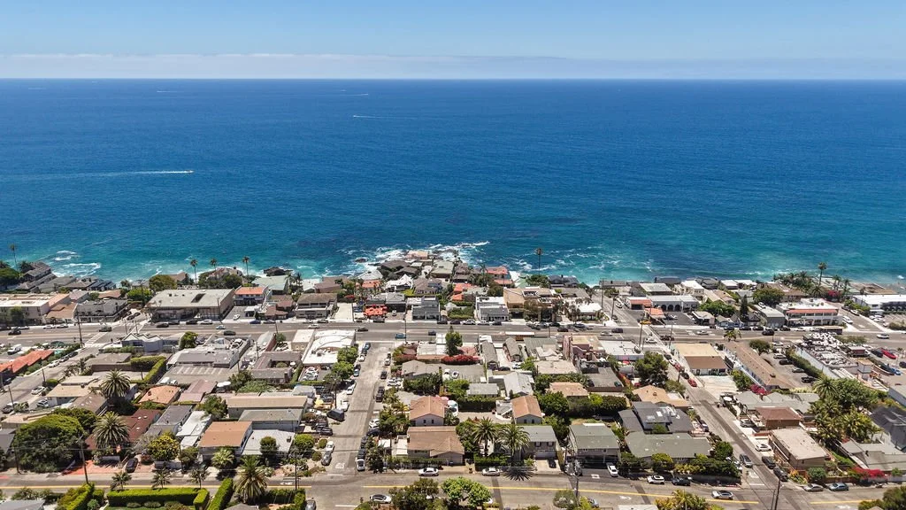 Aerial view of a coastal neighborhood with houses and streets in front of a large ocean, under a clear blue sky.